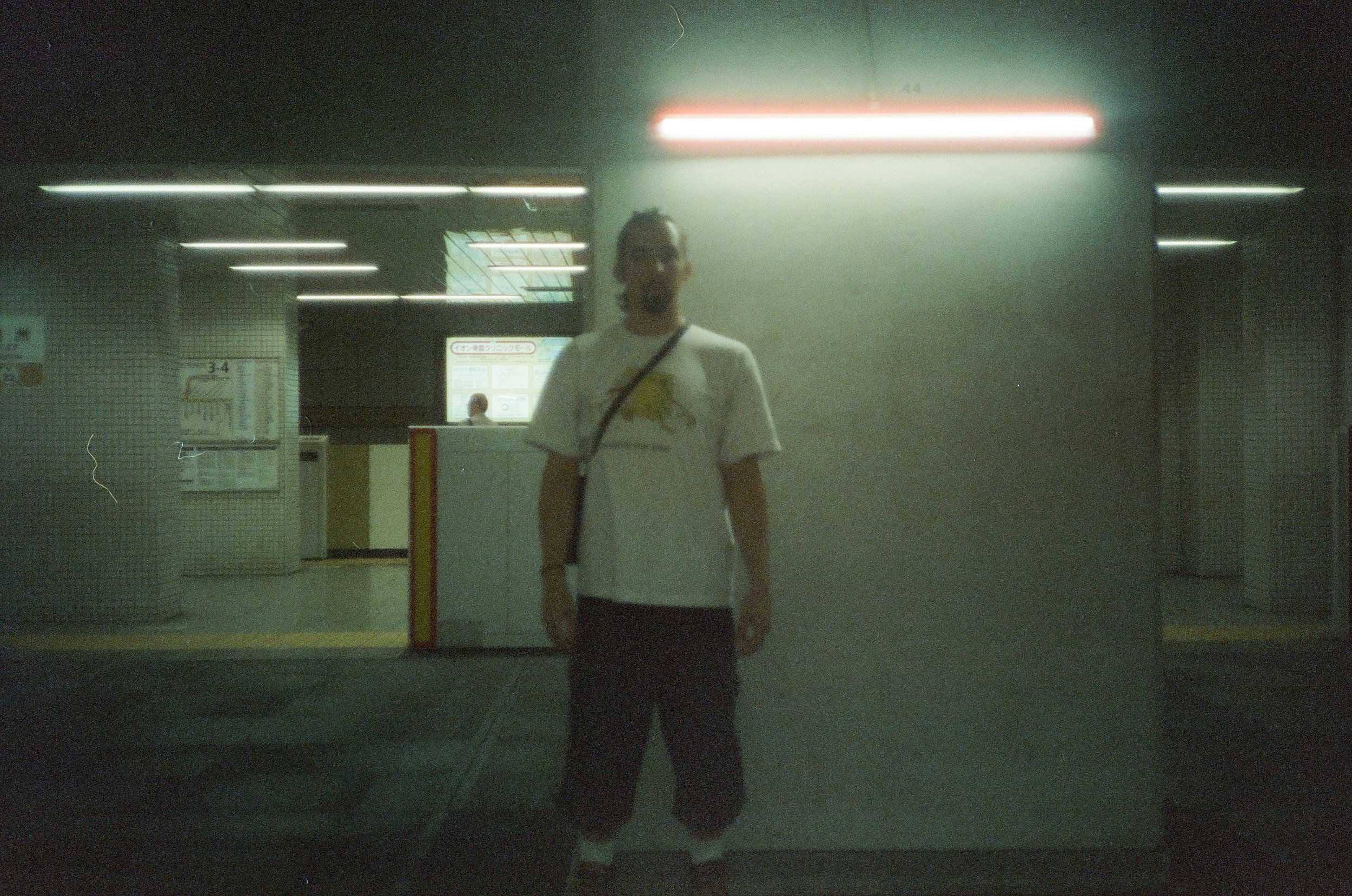 A person stands in a dimly lit underground parking garage or subway station, facing the camera, wearing a white T-shirt with a graphic, dark pants, and a crossbody bag, with fluorescent lights above and a ticket or information booth in the background