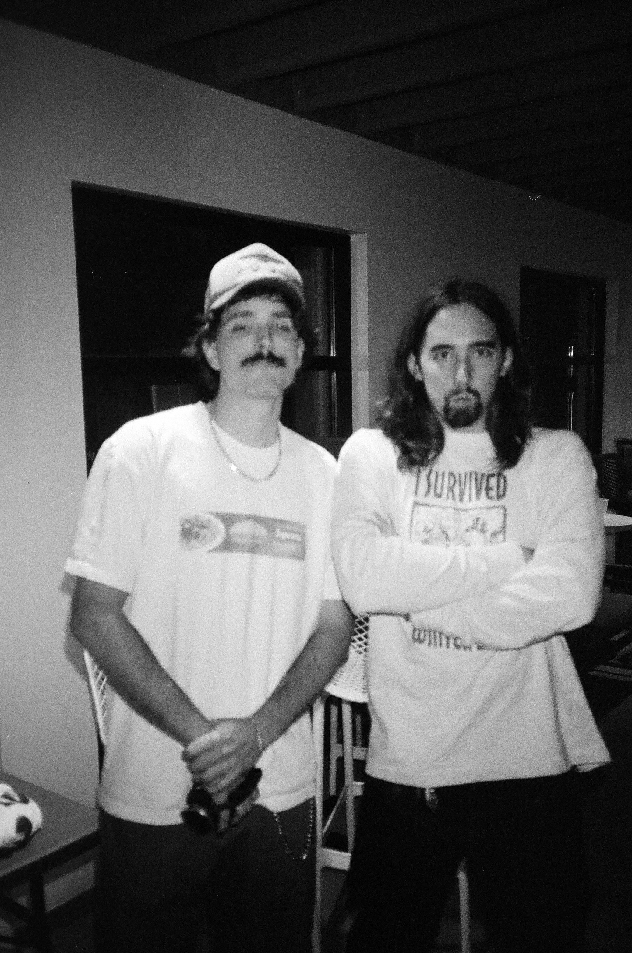 Two men standing indoors, one with a mustache and hat, the other with long hair and crossed arms, both wearing casual T-shirts.