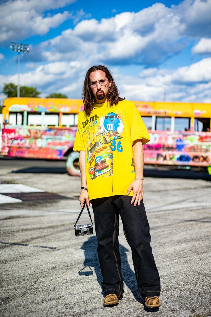 A man with long hair, glasses, and a beard stands outdoors on an empty parking lot during the day. He is wearing a bright yellow T-shirt featuring a racing cartoon design, black pants, and tan shoes. He holds a vintage camera with a strap in his righ