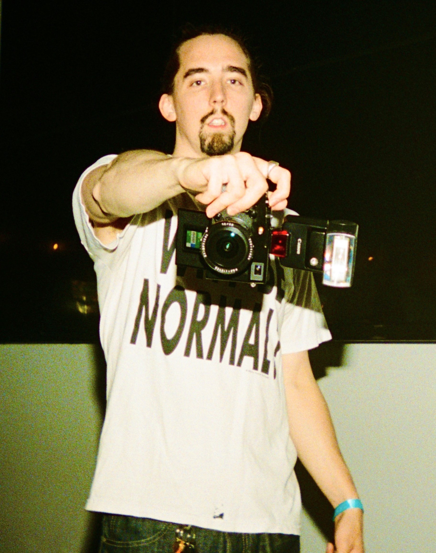 A man with long dark hair, a goatee, and a mustache holds a camera pointed at the viewer. He wears a white T-shirt with the word 'NORMAL' printed on it and a blue wristband, standing against a dark background.