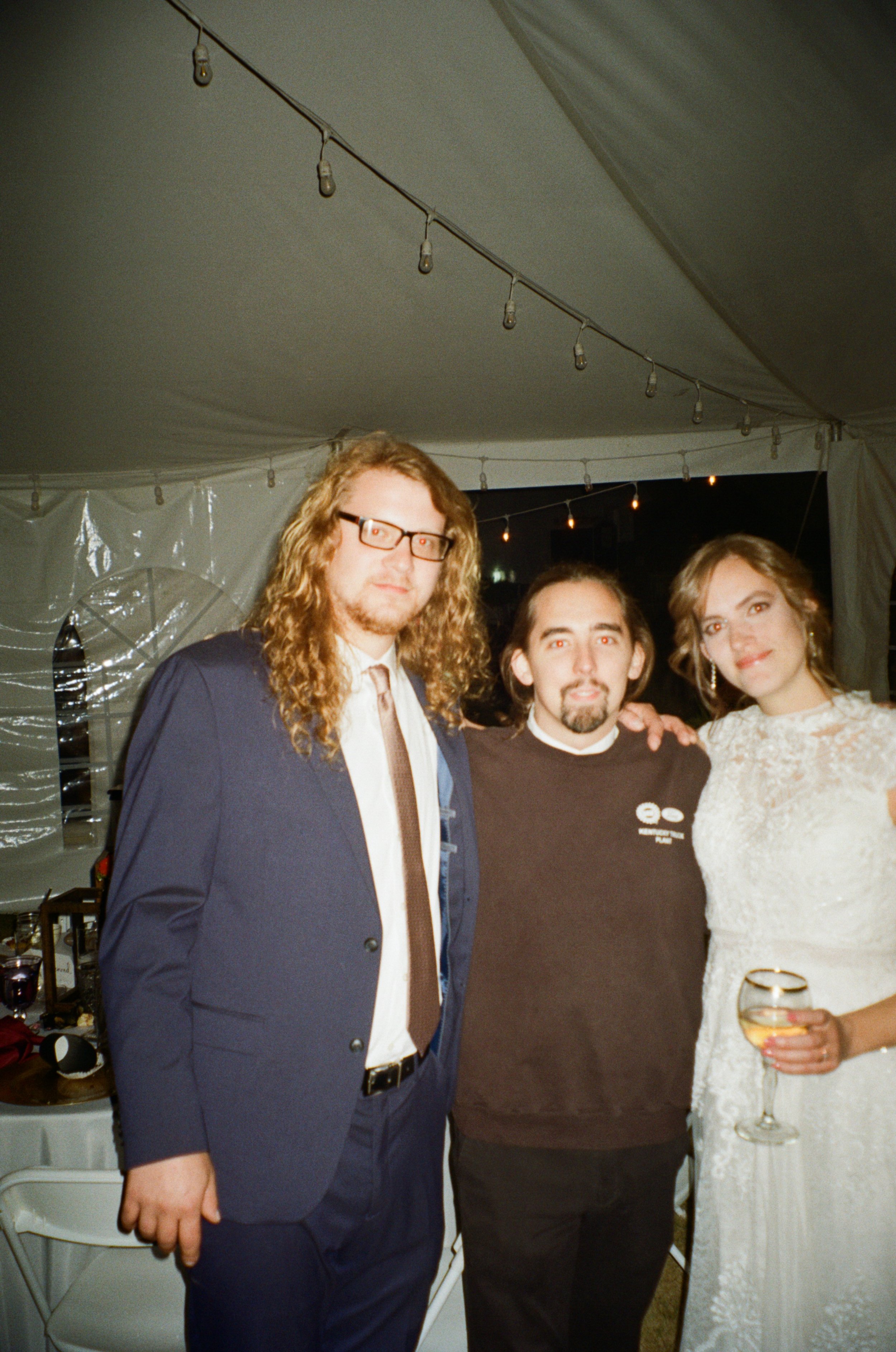 Three people smiling at a nighttime outdoor event, with two men and one woman, all dressed semi-formally. The woman on the right is holding a glass of wine.