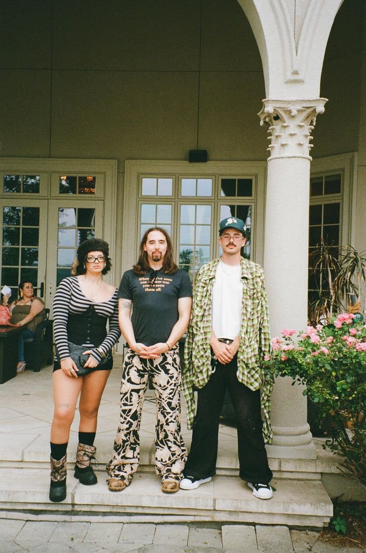 Three people standing on a porch in front of a house with large windows and a column, with two women sitting behind them and pink flowers on the right.
