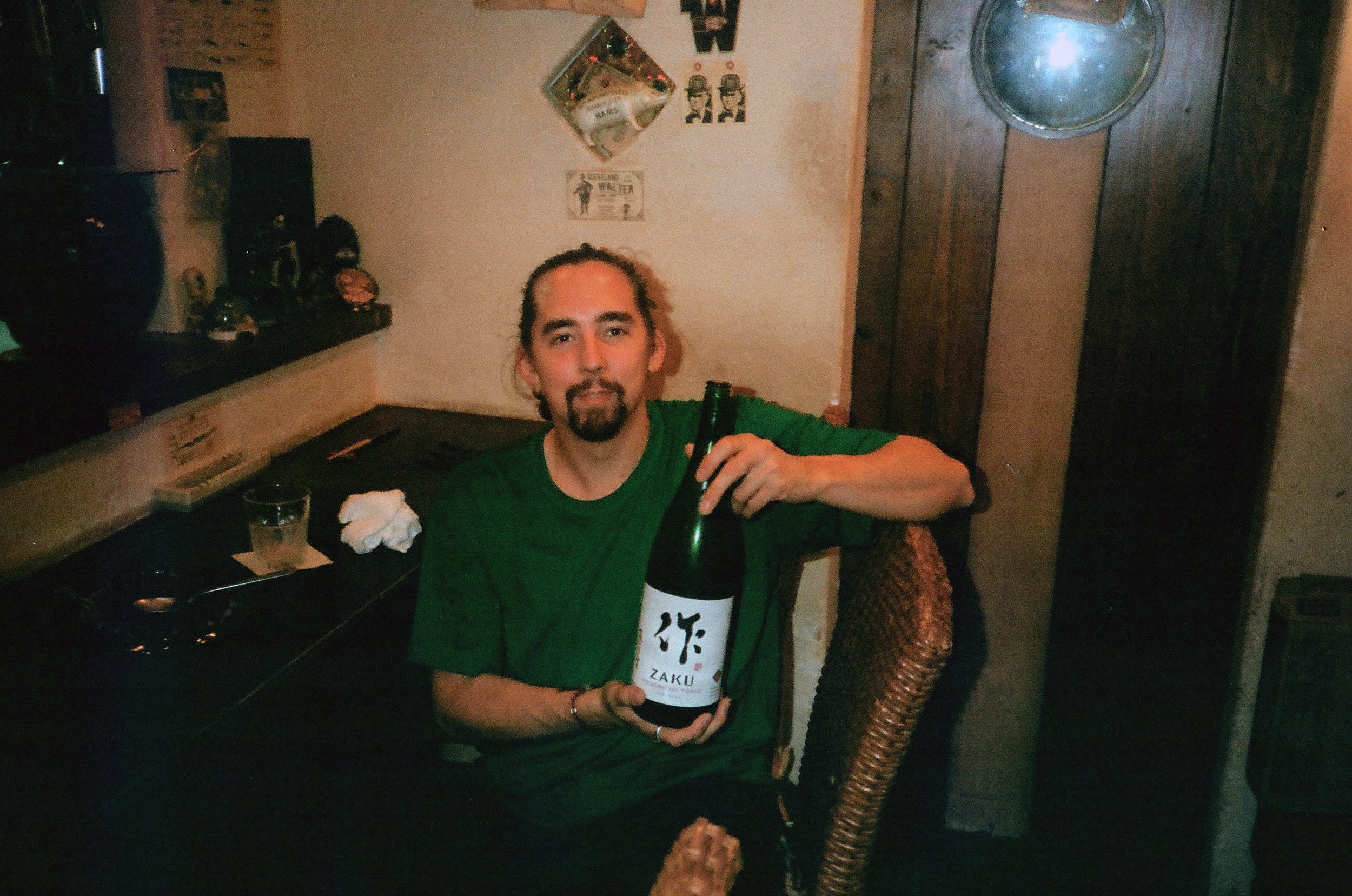 A man with long hair and a goatee sitting at a table, holding a bottle with a label written in Japanese characters. He is wearing a green shirt and is in a room decorated with various pictures and items on the wall.