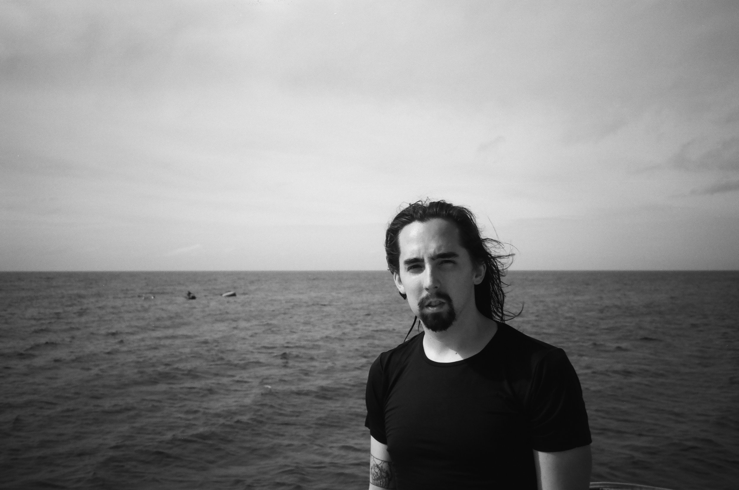 A man with long hair and a goatee stands at the edge of a body of water, looking at the camera with a serious expression in a black and white photo.