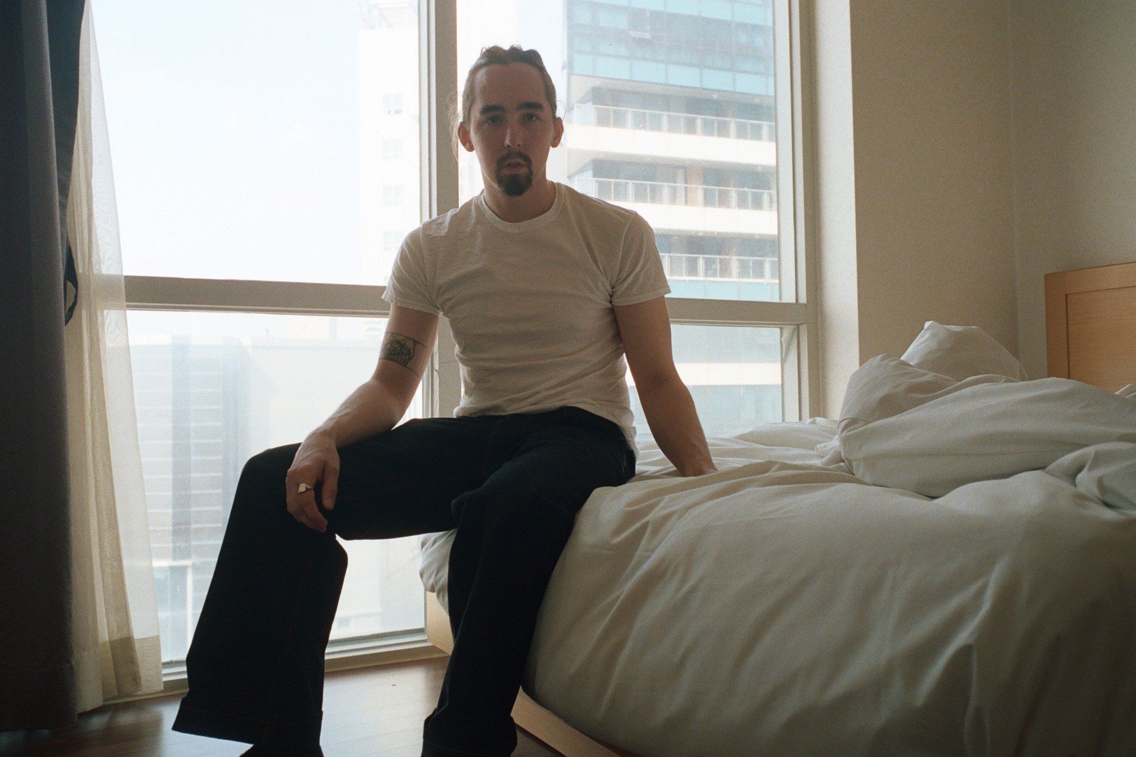 A man with a goatee and hairstyle sitting on the edge of a bed in a room with large windows showing tall buildings outside, wearing a white t-shirt and black pants.