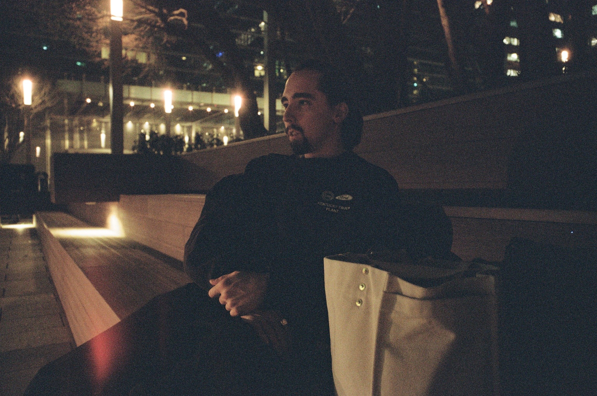 A man with dark hair and a beard sitting on a wooden bench at night, wearing a black uniform with a logo that says 'Kendrick Truck Plant.' He is looking off to the side, with city lights and a modern building in the background.