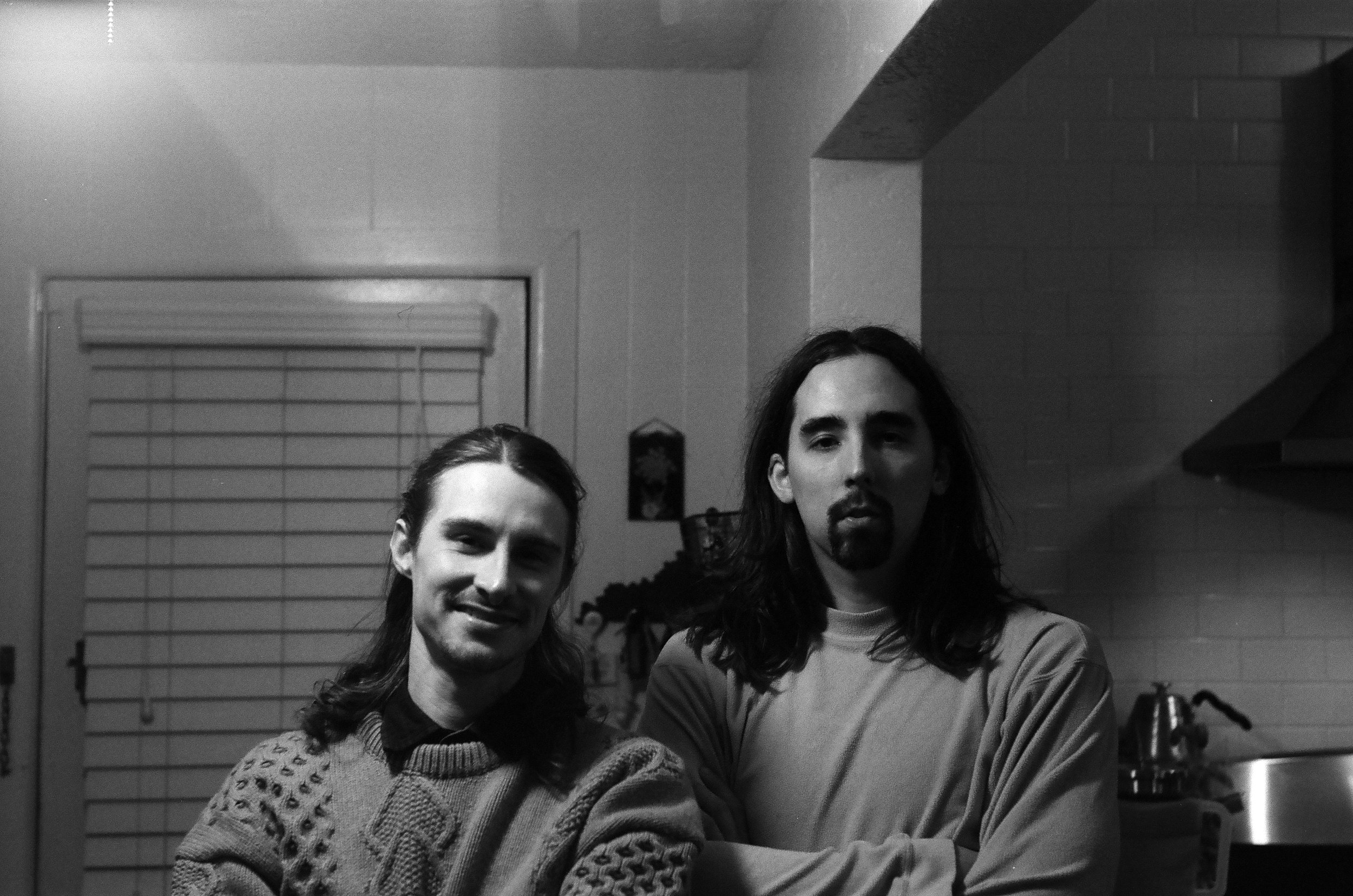 Two men with long hair, one smiling and the other with a serious expression, standing in a kitchen.