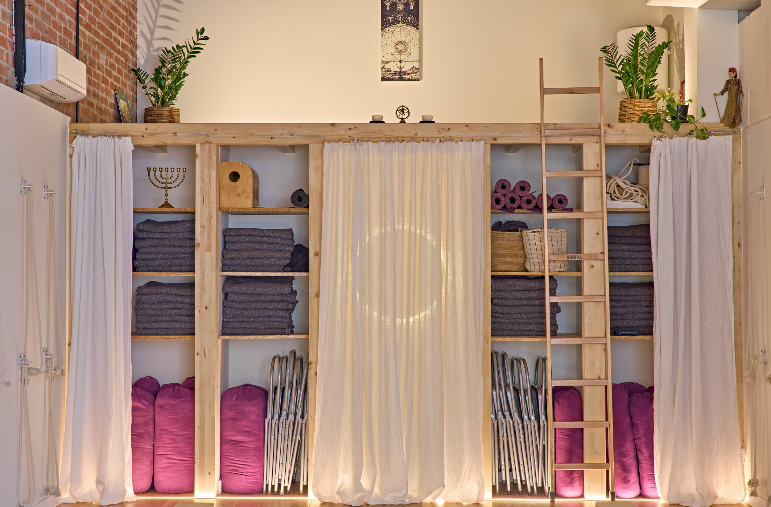 Sala de espera con plantas, sillas metálicas y de madera, una mesa baja de madera, alfombra blanca, reloj de pared, luminarias de papel, y cortinas blancas.