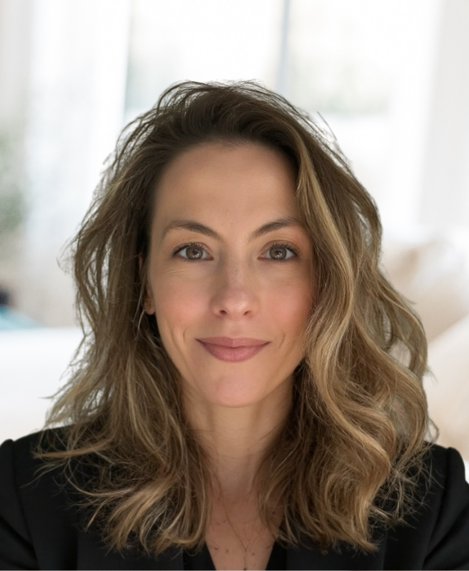 Dr Sara Sherif with wavy, shoulder-length brown hair and light skin, smiling softly at the camera in a well-lit indoor setting.