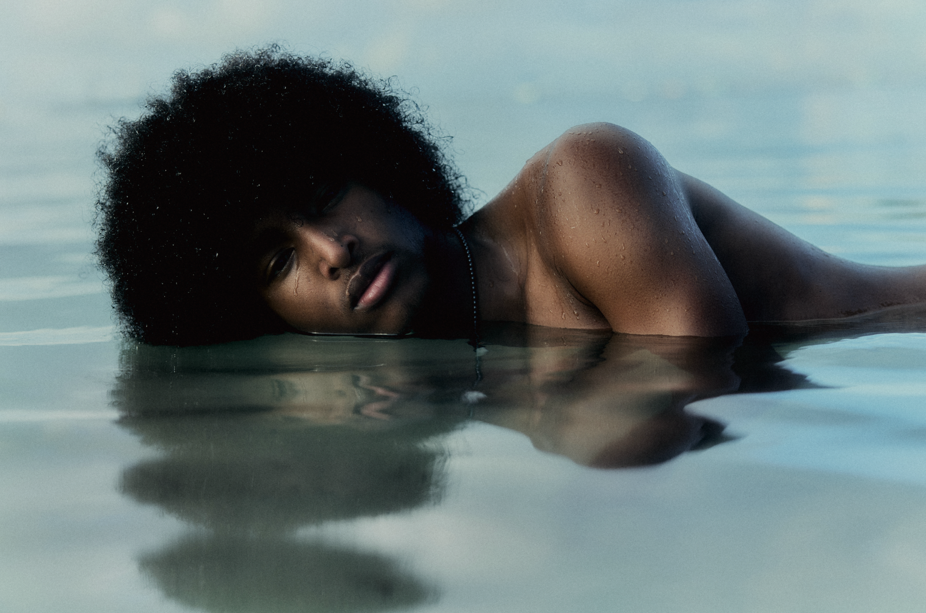 A male in water against a reflective surface with her upper body exposed; water droplets are visible on her skin.