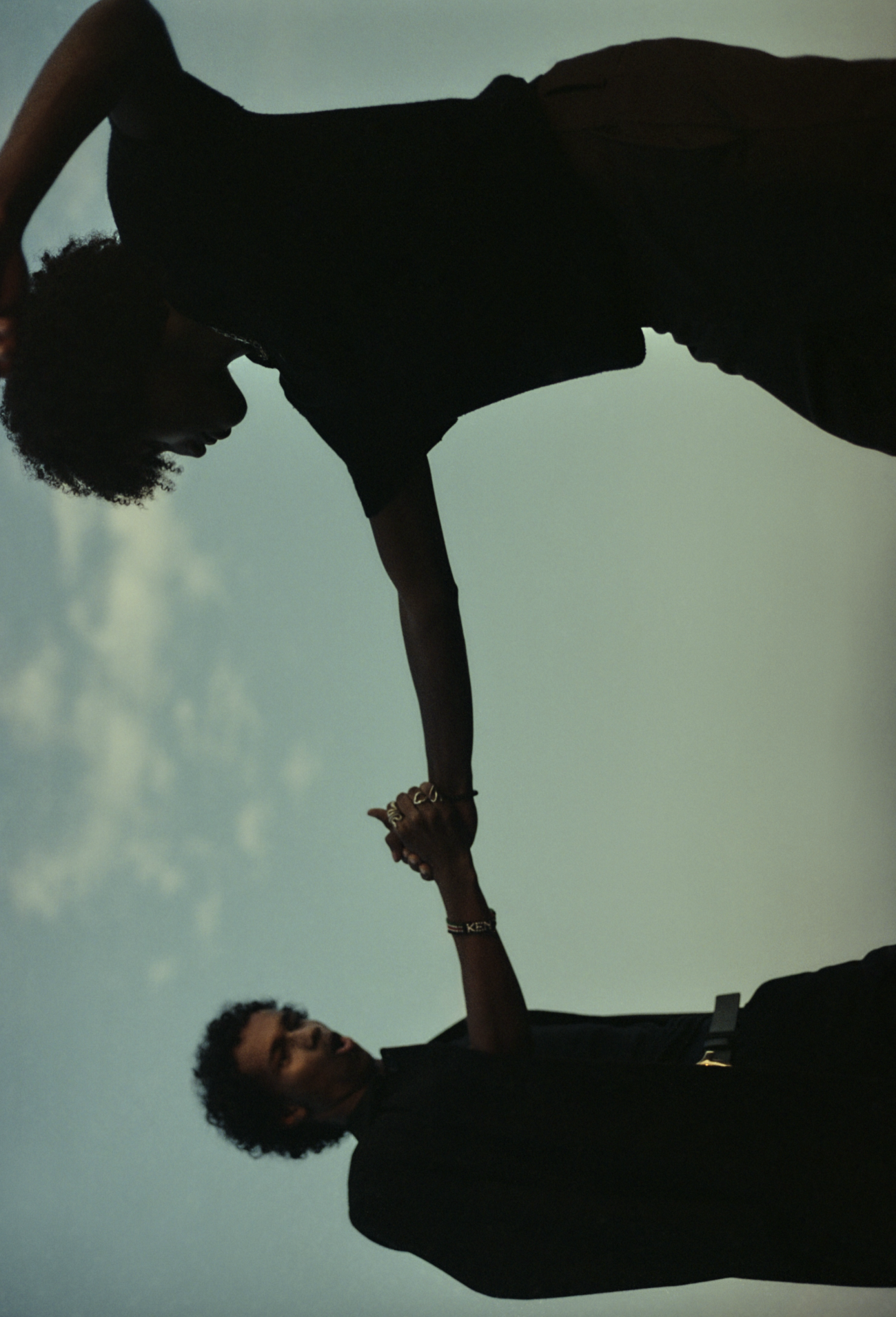 Two people with curly hair and glasses holding hands, silhouetted against a light sky during likely sunset or sunrise.