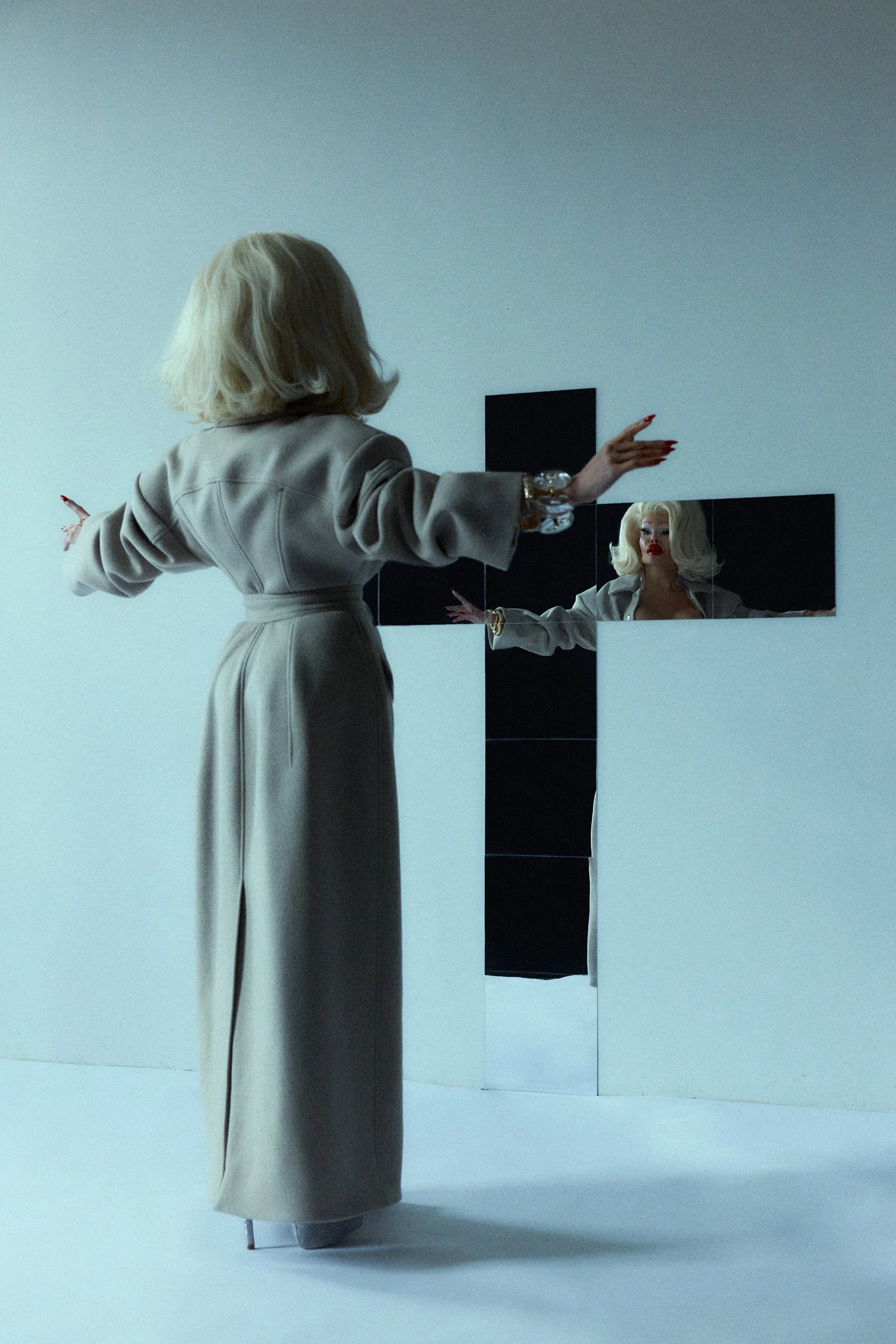 Amanda Lepore with blonde hair and red lipstick standing in front of a gallery wall with three mirrors reflecting her face and arms, wearing a long beige coat with her arms outstretched.