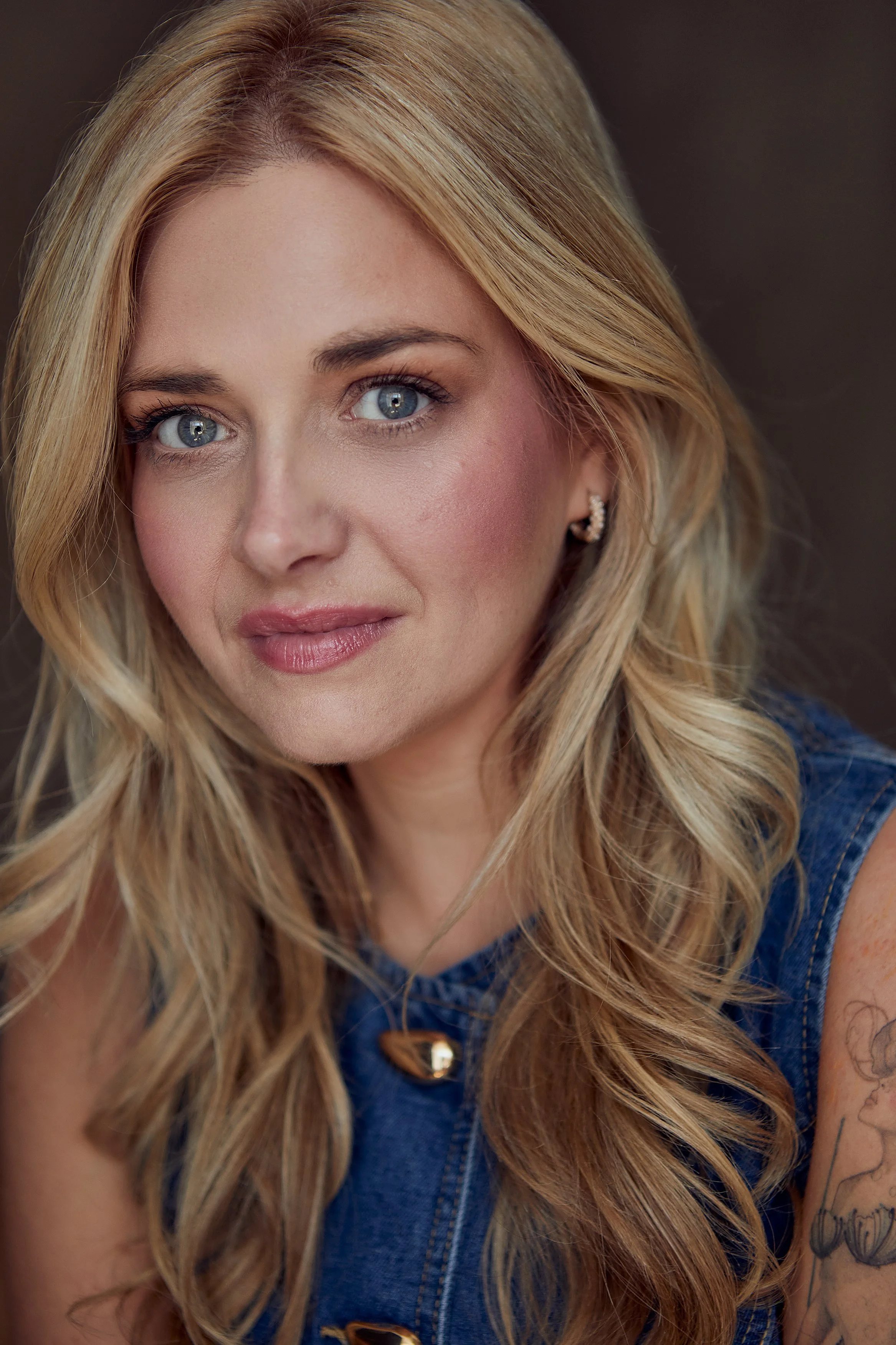 Close-up of a woman with blonde hair, blue eyes, wearing a denim top and earrings, with tattoos on her arm.
