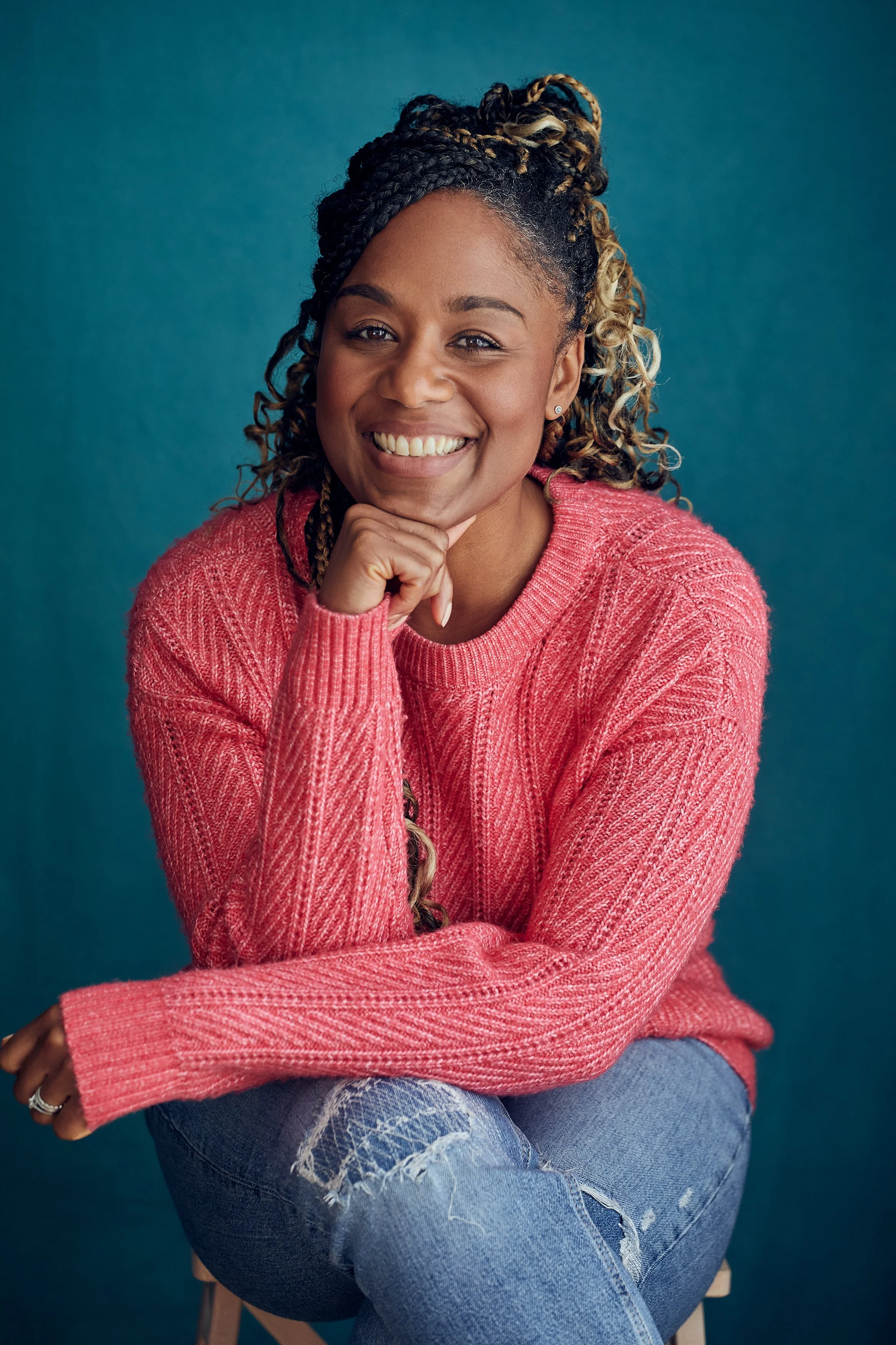 A woman with curly hair in a braid styled up, wearing a pink sweater and ripped jeans, sitting with her chin resting on her hand and smiling against a blue background.