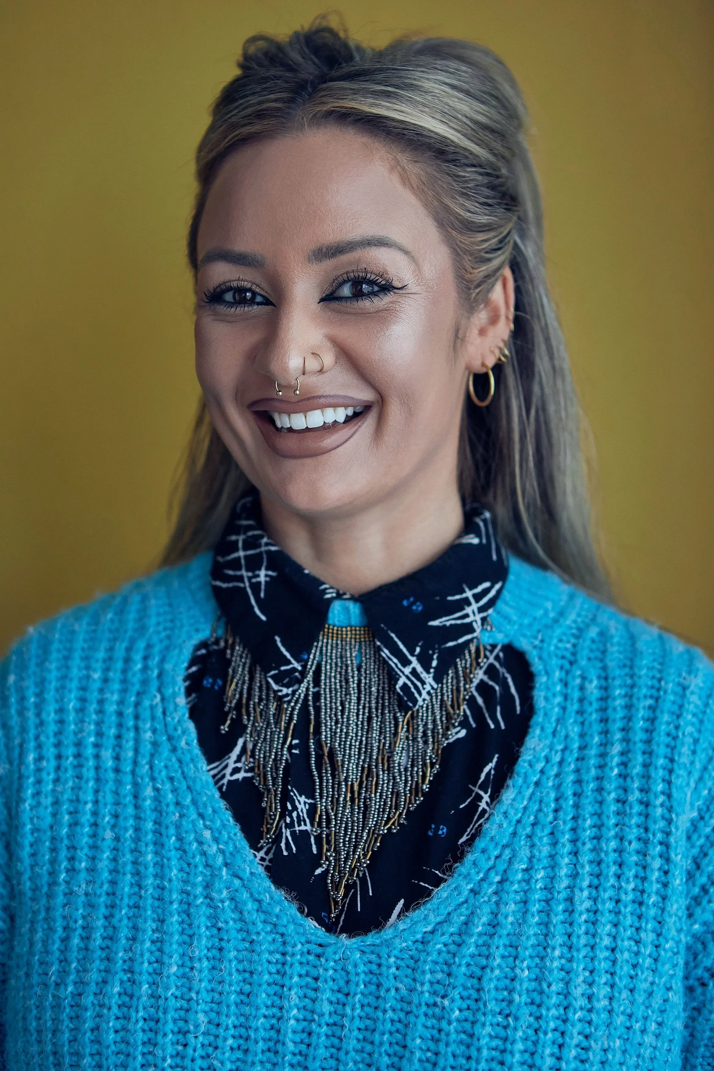A woman with light skin, blonde hair, and blue eyes smiling, wearing a black patterned shirt, a blue sweater, and layered necklaces, standing against a yellow background.