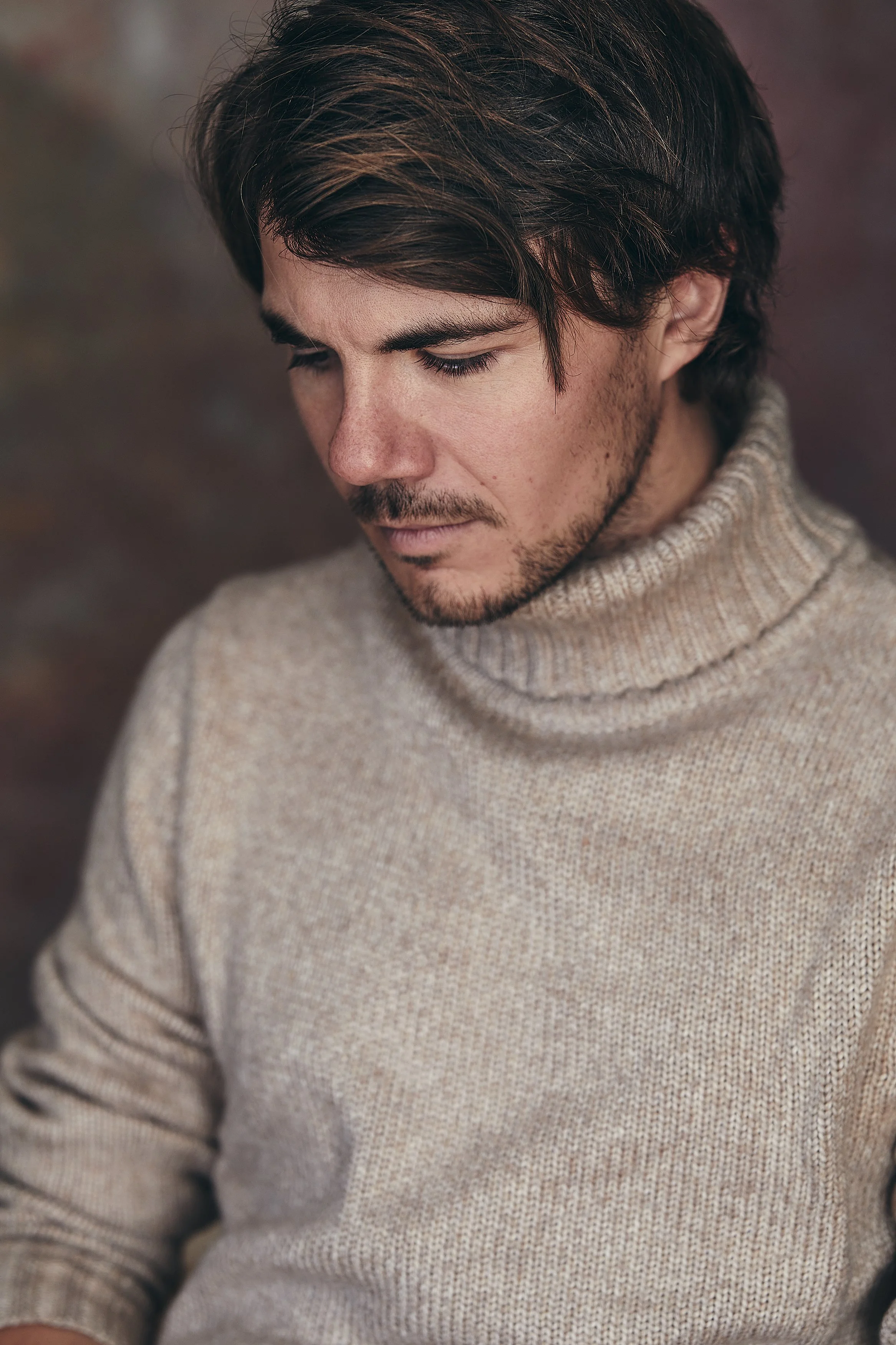 A young man with dark hair and a light beard, wearing a beige knit turtleneck sweater, looking down with a contemplative expression.