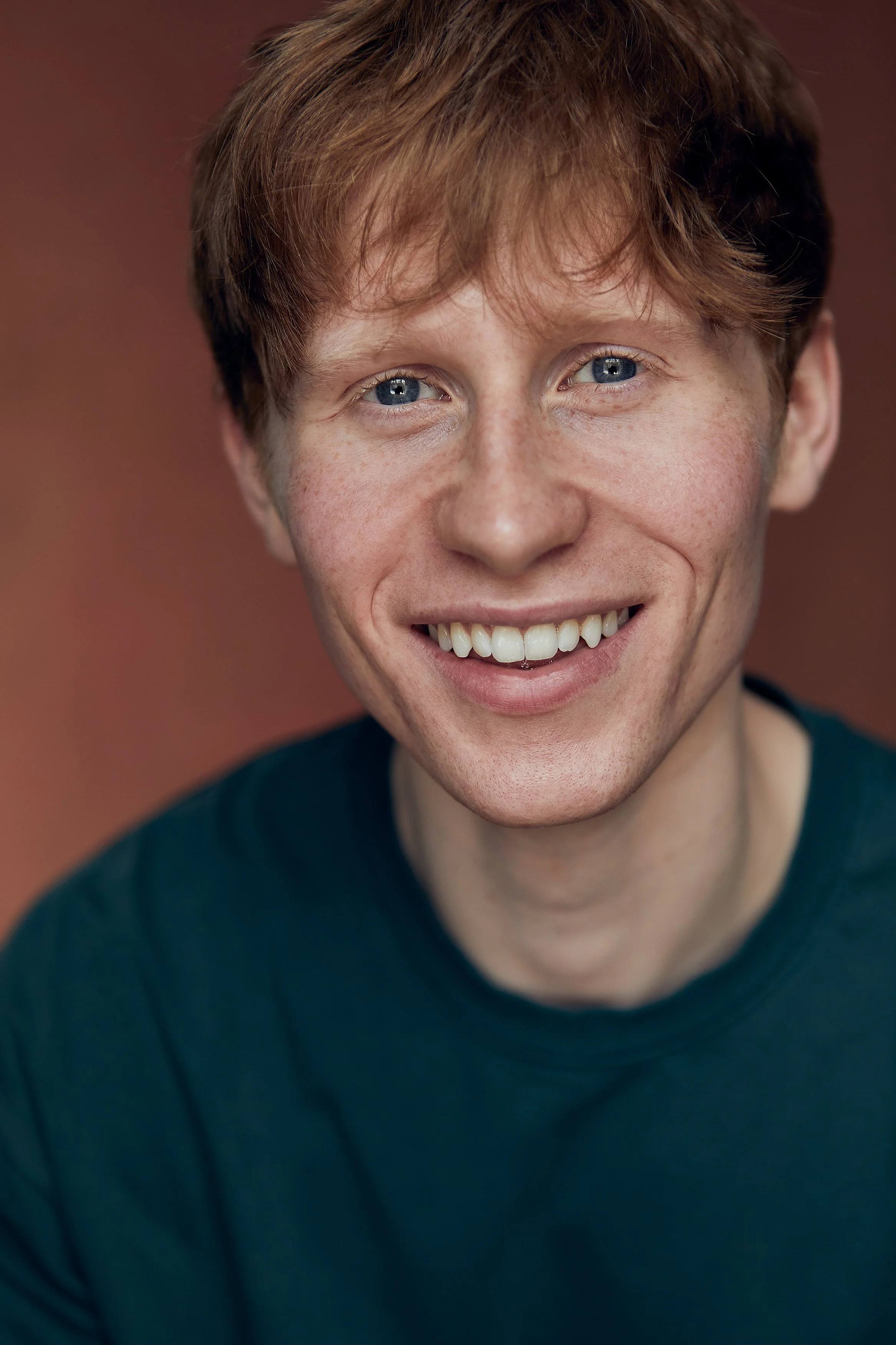 Close-up of a young person with short red hair, blue eyes, freckles, smiling, wearing a dark green top.