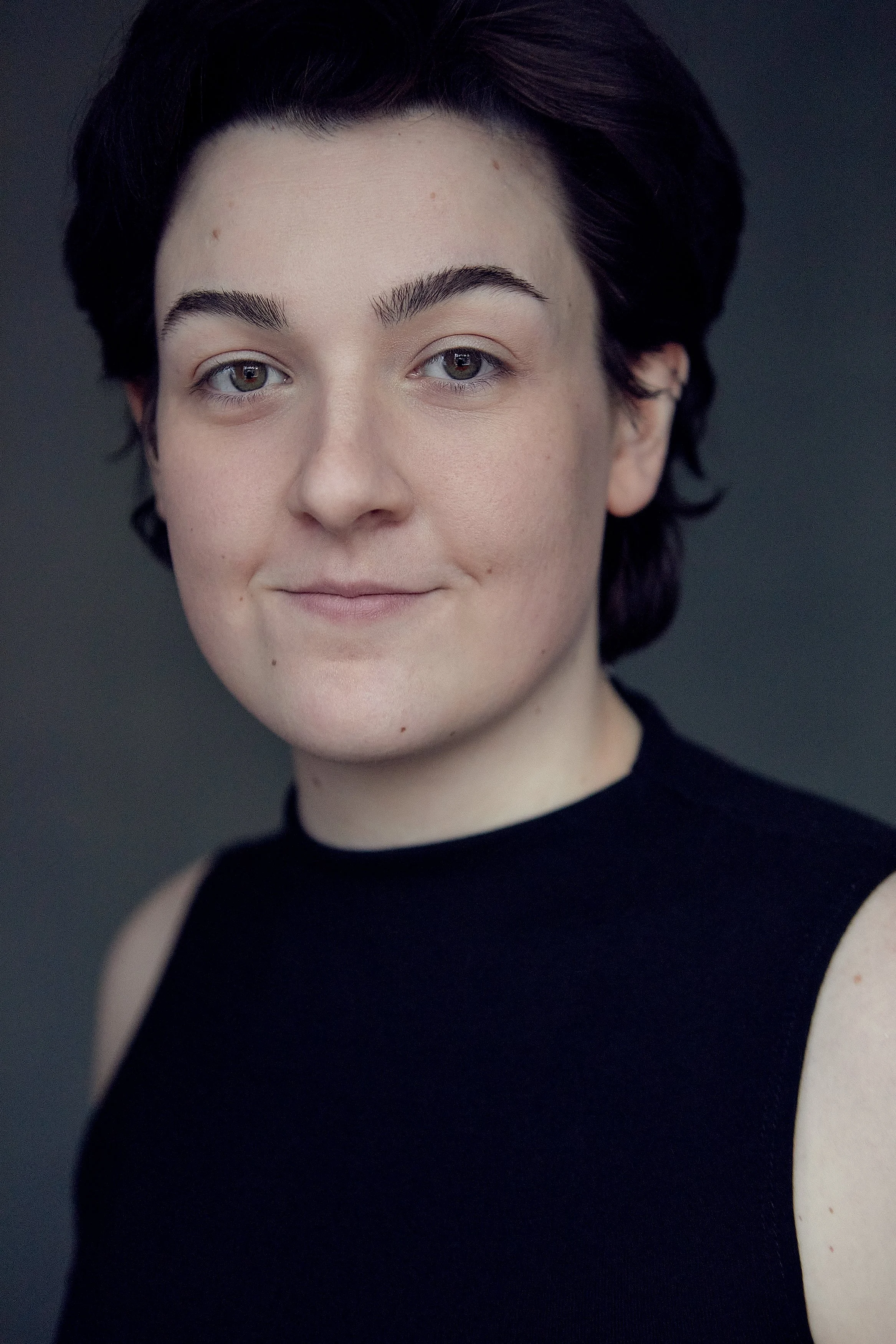 Close-up portrait of a young person with short dark hair, wearing a black sleeveless top, looking at the camera with a slight smile against a dark background.
