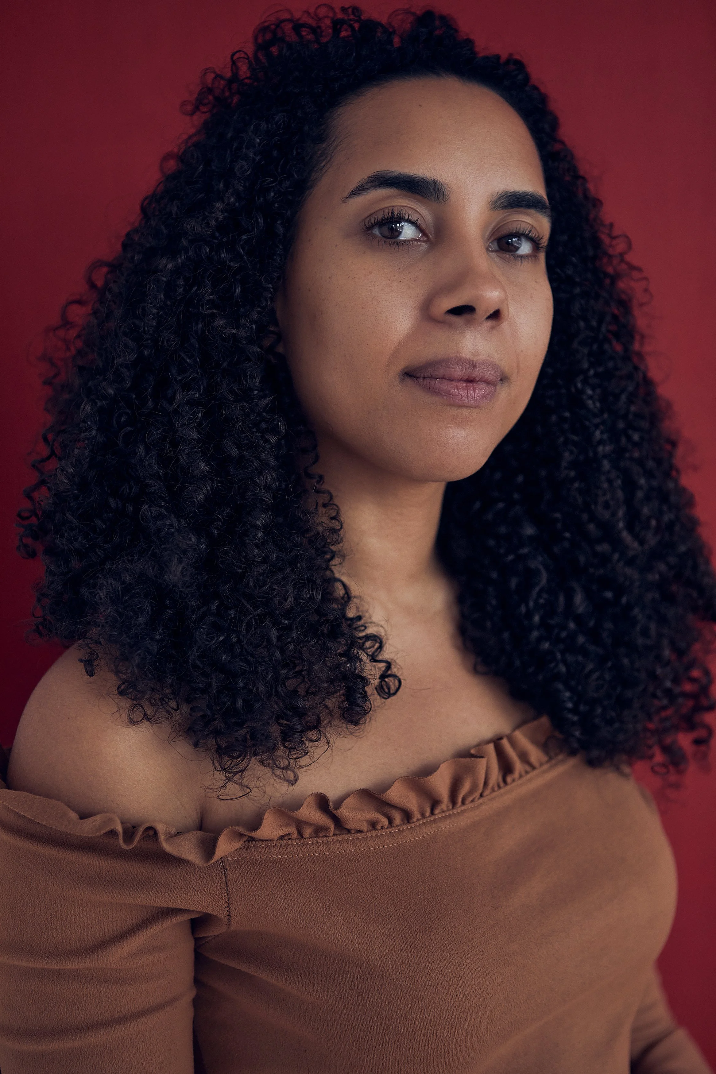 A woman with long, curly dark hair and a medium skin tone looks confidently at the camera against a red background, wearing a brown off-the-shoulder top.