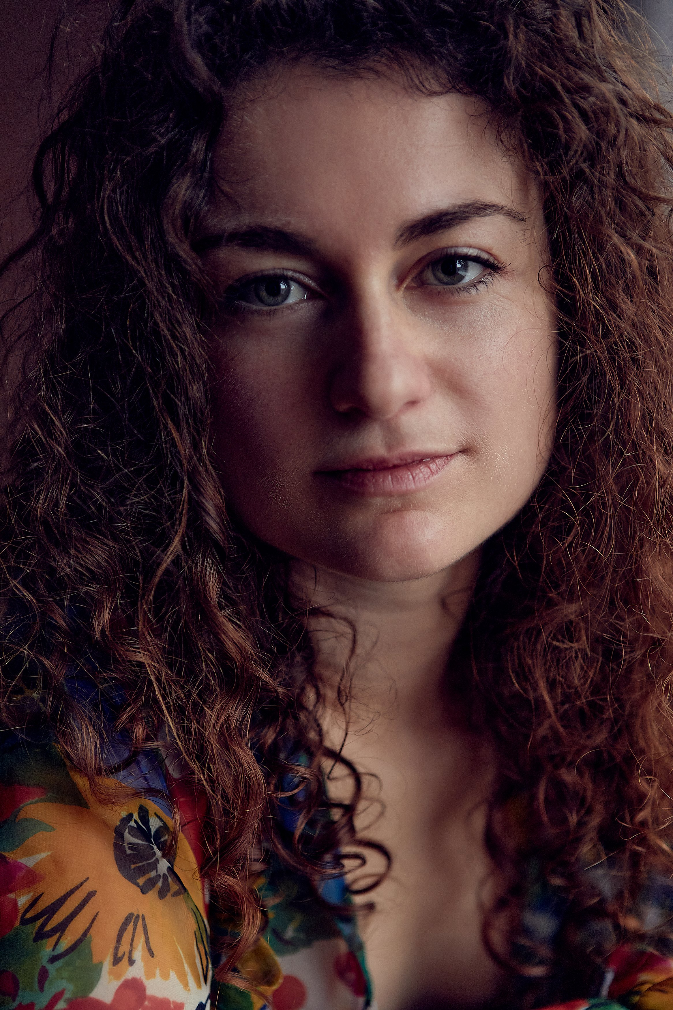 Close-up of a woman with curly brown hair and blue eyes, wearing a colorful floral top.