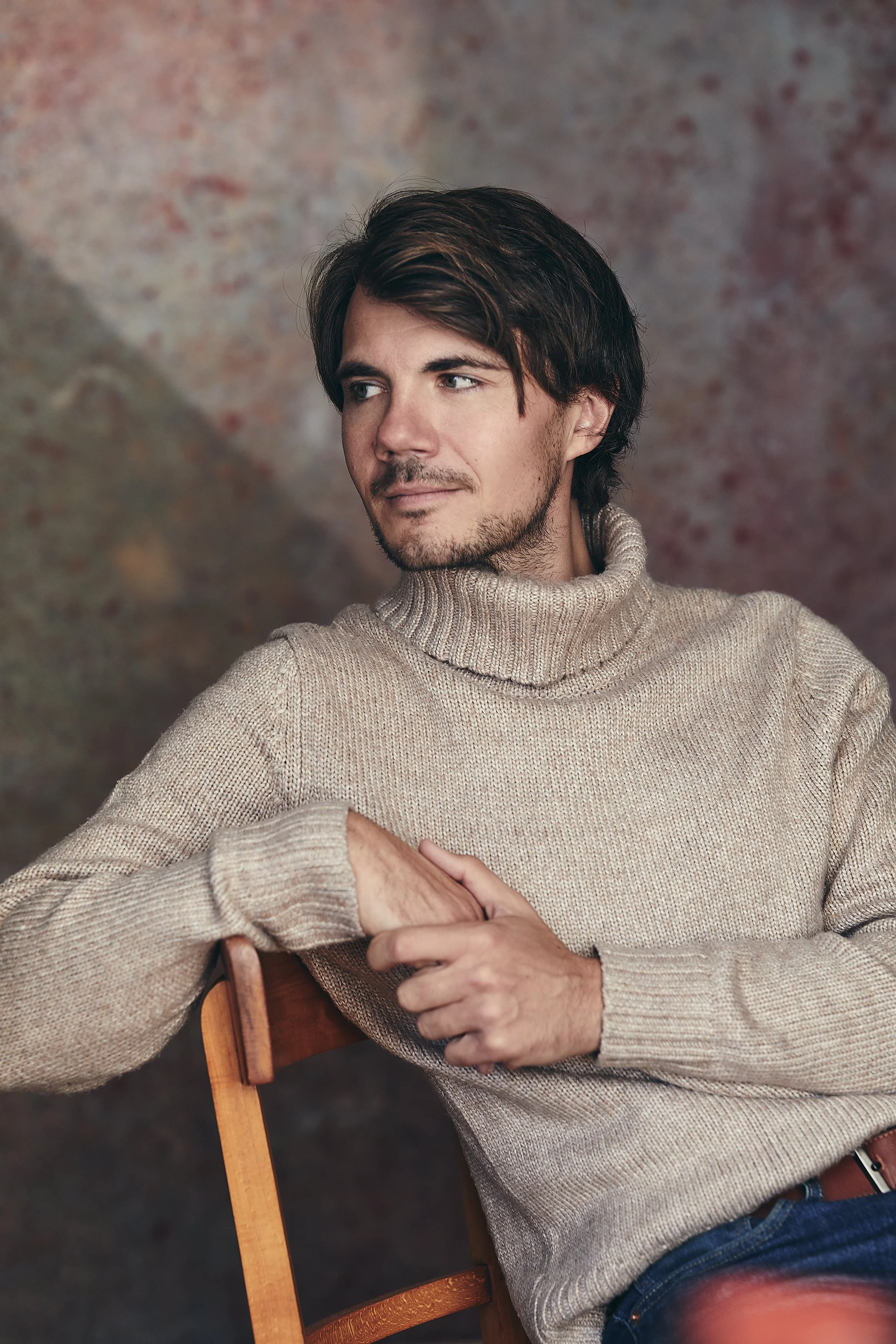 A young man with dark hair and a beard sitting on a wooden chair, wearing a beige turtleneck sweater, looking to his left with a contemplative expression.