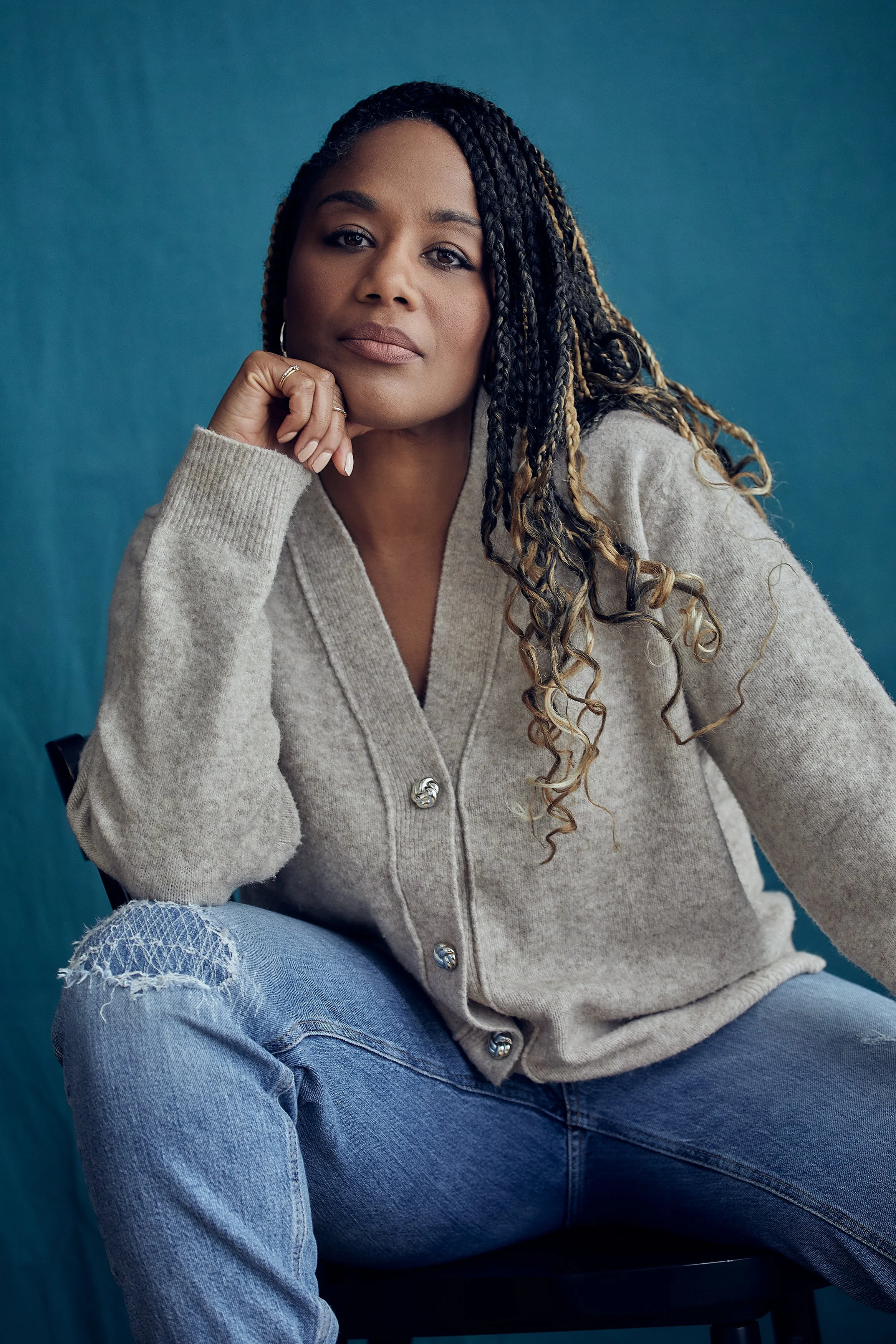 A woman with dark braided hair and tan skin sitting against a blue background. She is wearing a beige cardigan with buttons and ripped light blue jeans, resting her chin on her hand and looking confidently at the camera.
