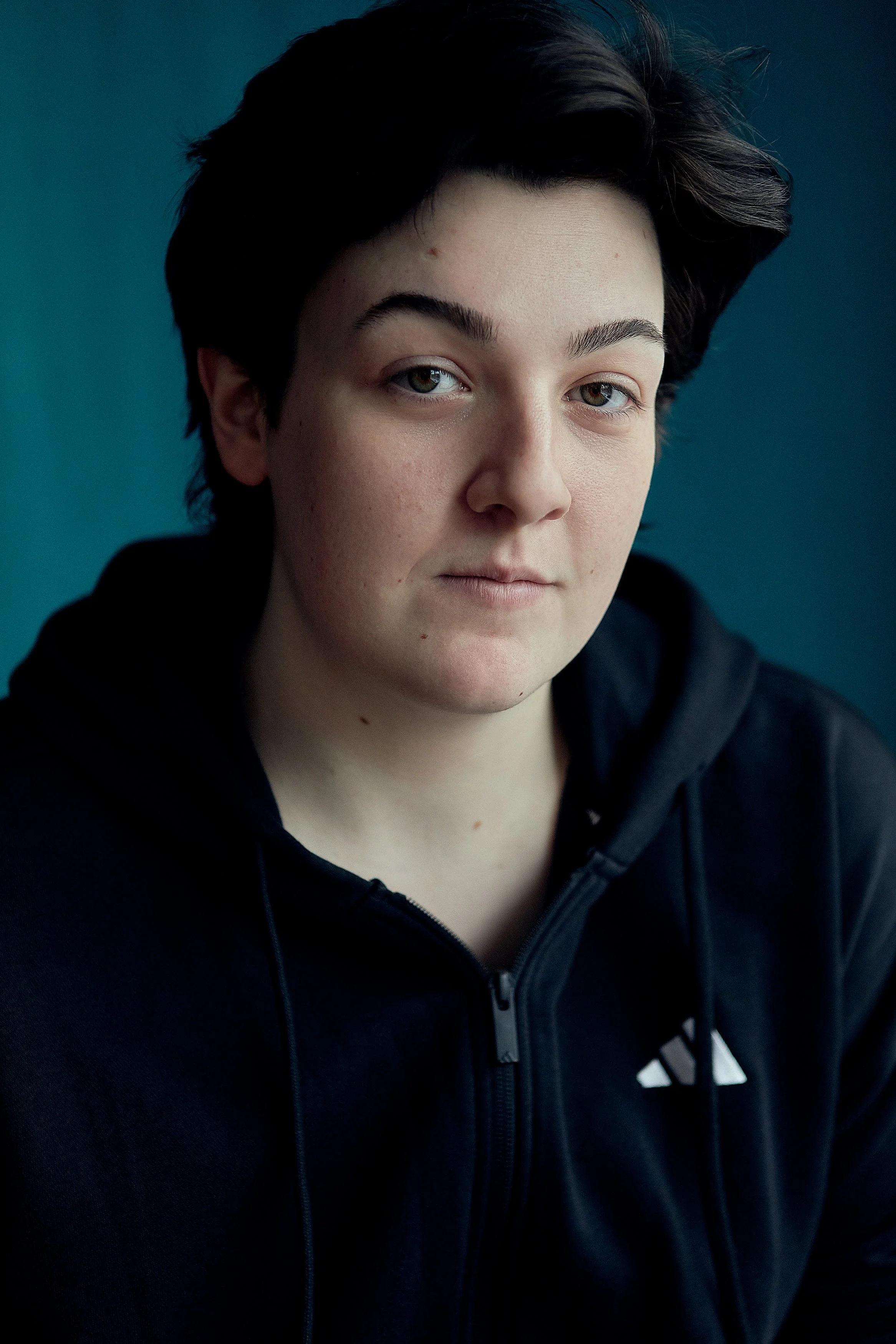 actor headshots liverpool - A young person with short dark hair and light skin wearing a black hoodie, looking directly at the camera against a teal background.
