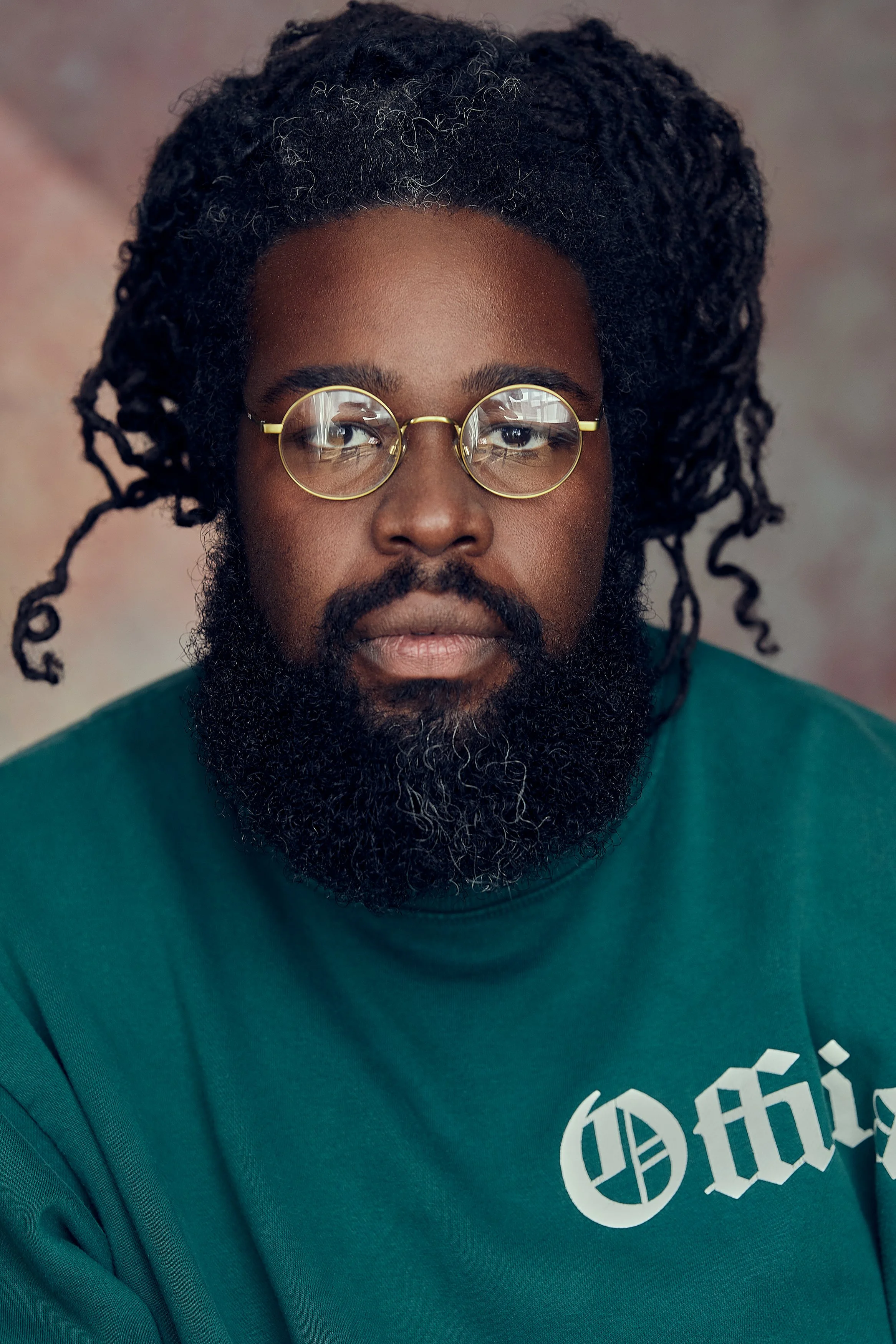 Portrait of an African American man with dreadlocks, a beard, and glasses, wearing a dark green sweatshirt with text on it, against a multicolored background.