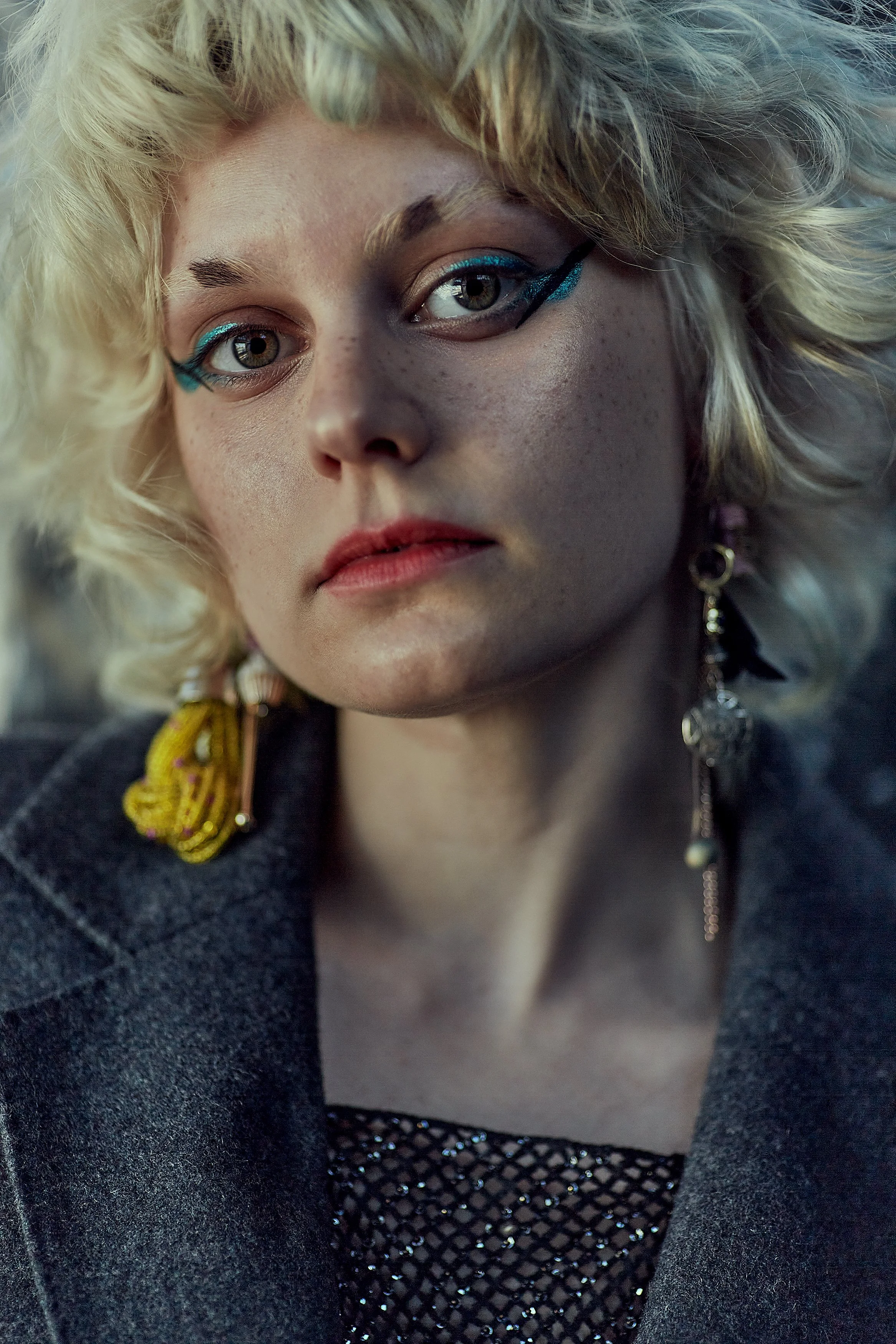 Close-up portrait of a woman with platinum blonde, curly hair wearing bold makeup, including blue eyeliner and shimmering eye makeup, with a confident expression. She is adorned with statement earrings and dressed in dark clothing.