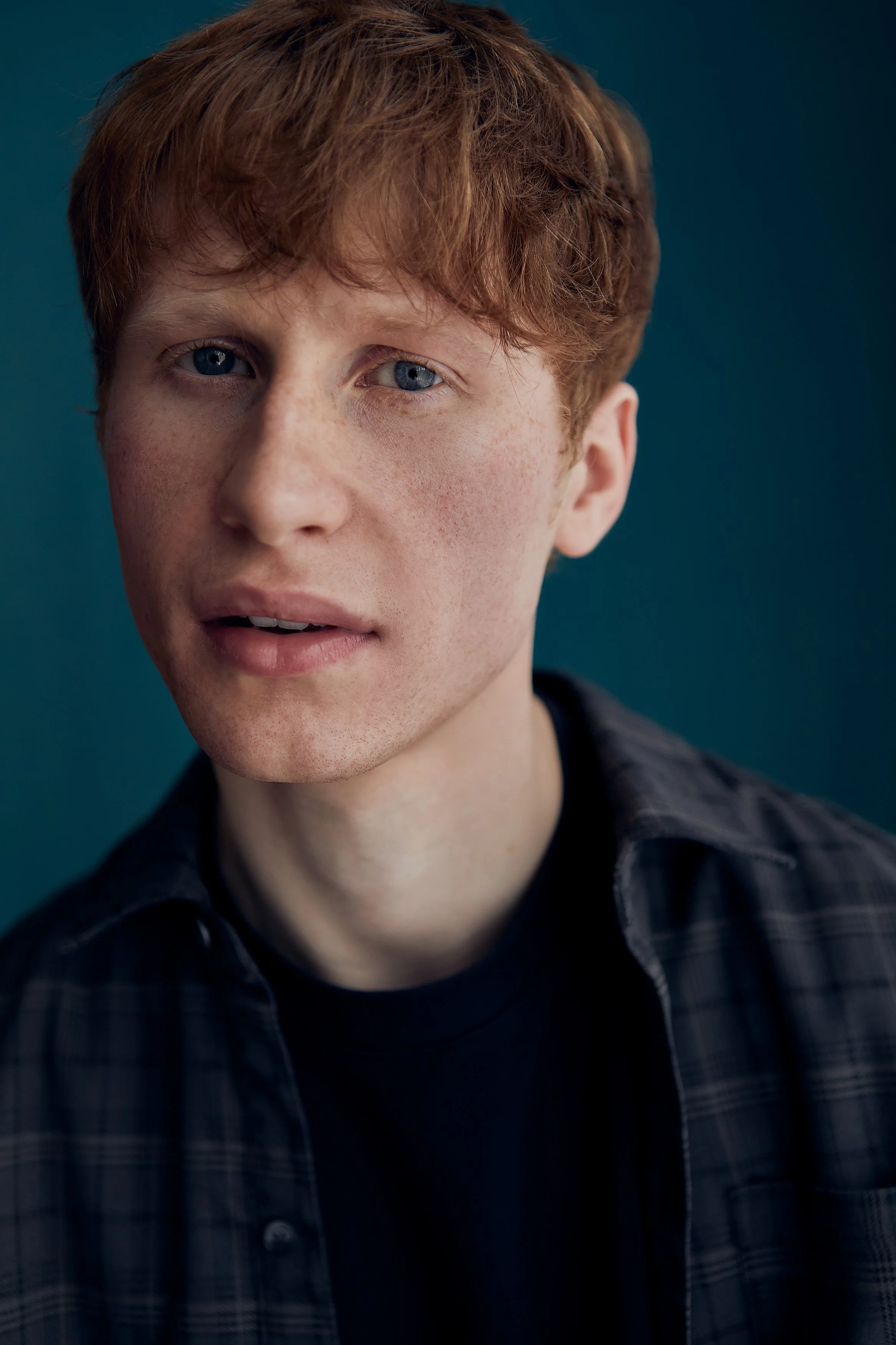 A close-up portrait of a young man with red hair and blue eyes, wearing a dark plaid shirt against a dark teal background.