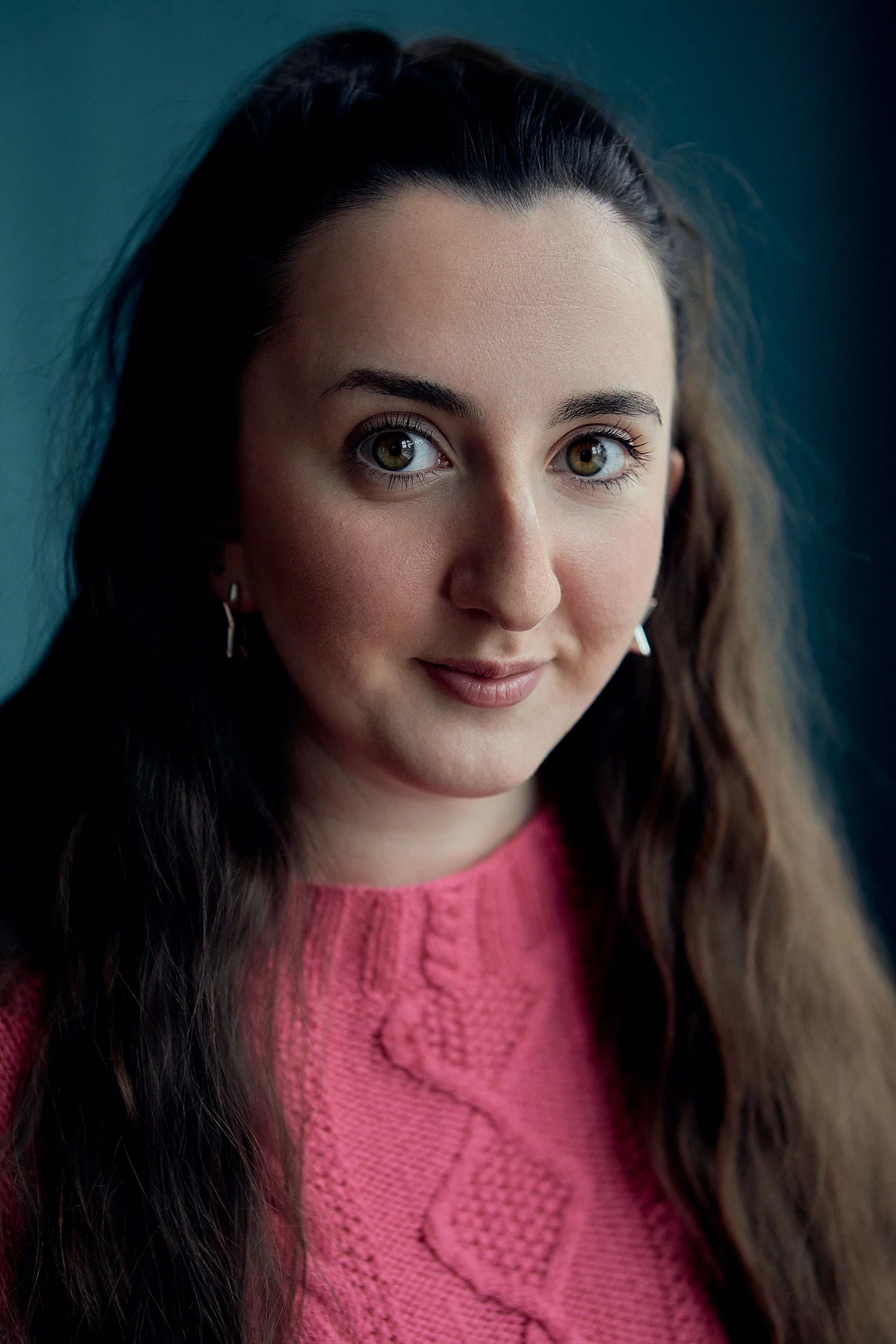 Close-up of a young woman with long, dark hair, wearing a pink sweater, smiling softly, with a teal background.