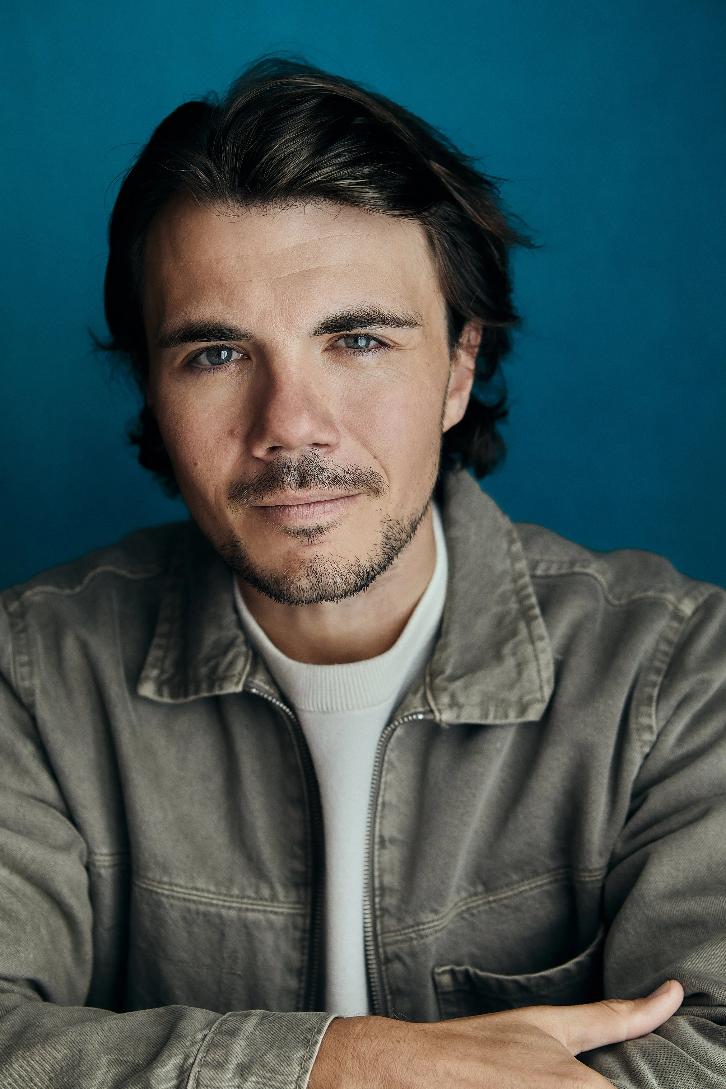 Close-up of a young man with dark hair, blue eyes, slight beard, wearing a grey jacket and white shirt, posing against a blue background.