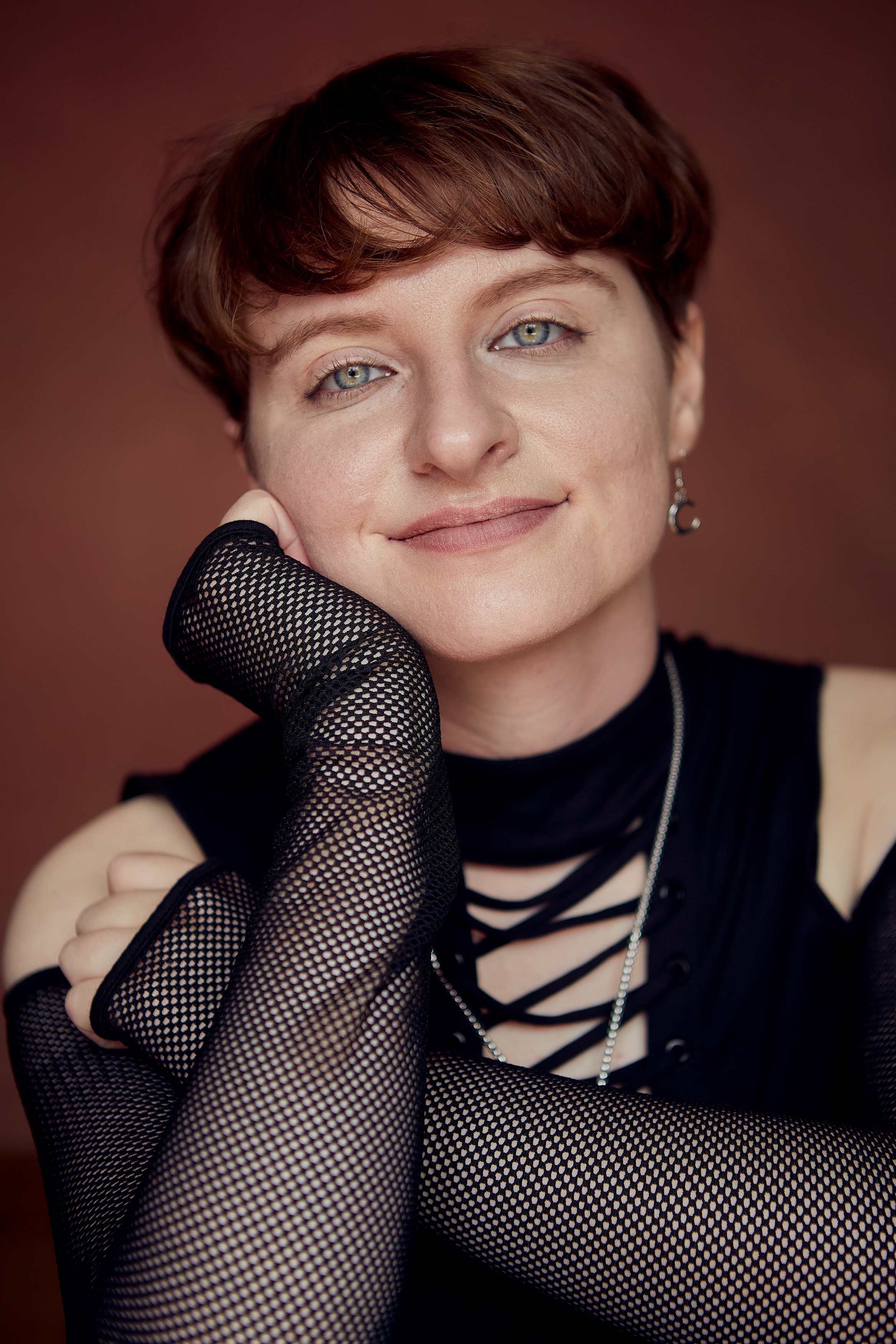 A woman with short red hair and blue eyes, wearing fishnet gloves and a black top with cut-out details, smiling gently at the camera with a warm brown background.