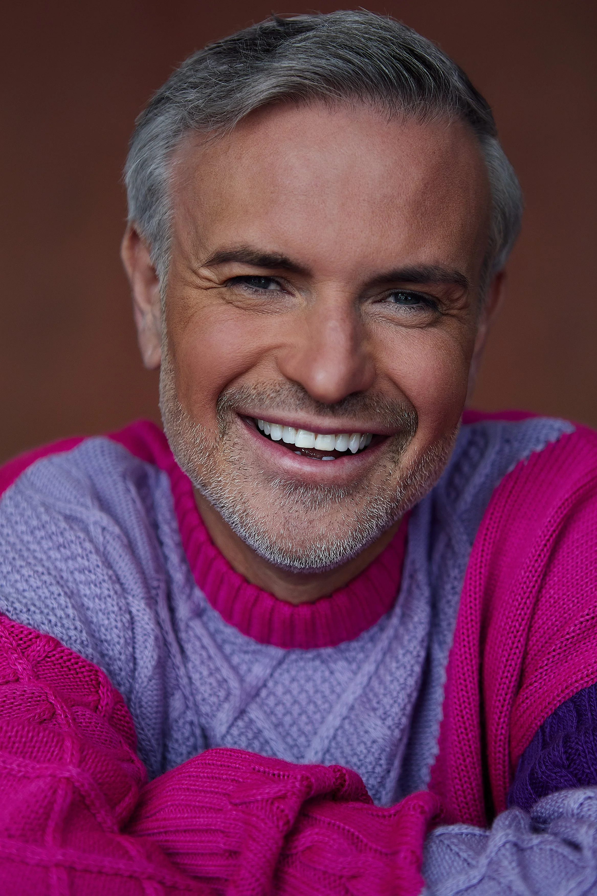 Close-up of a smiling man with gray hair and a beard, wearing a pink and purple knitted sweater.