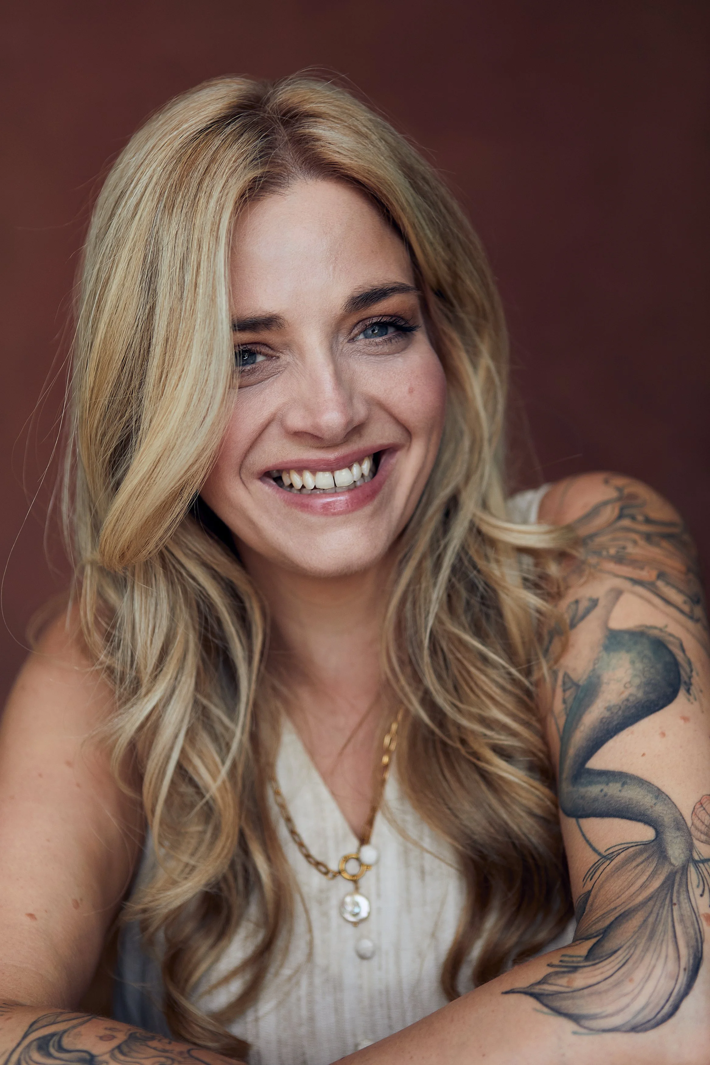 Close-up of a smiling woman with long blonde hair and tattoos on her arm, wearing a white sleeveless top and gold jewelry, positioned against a brown background.