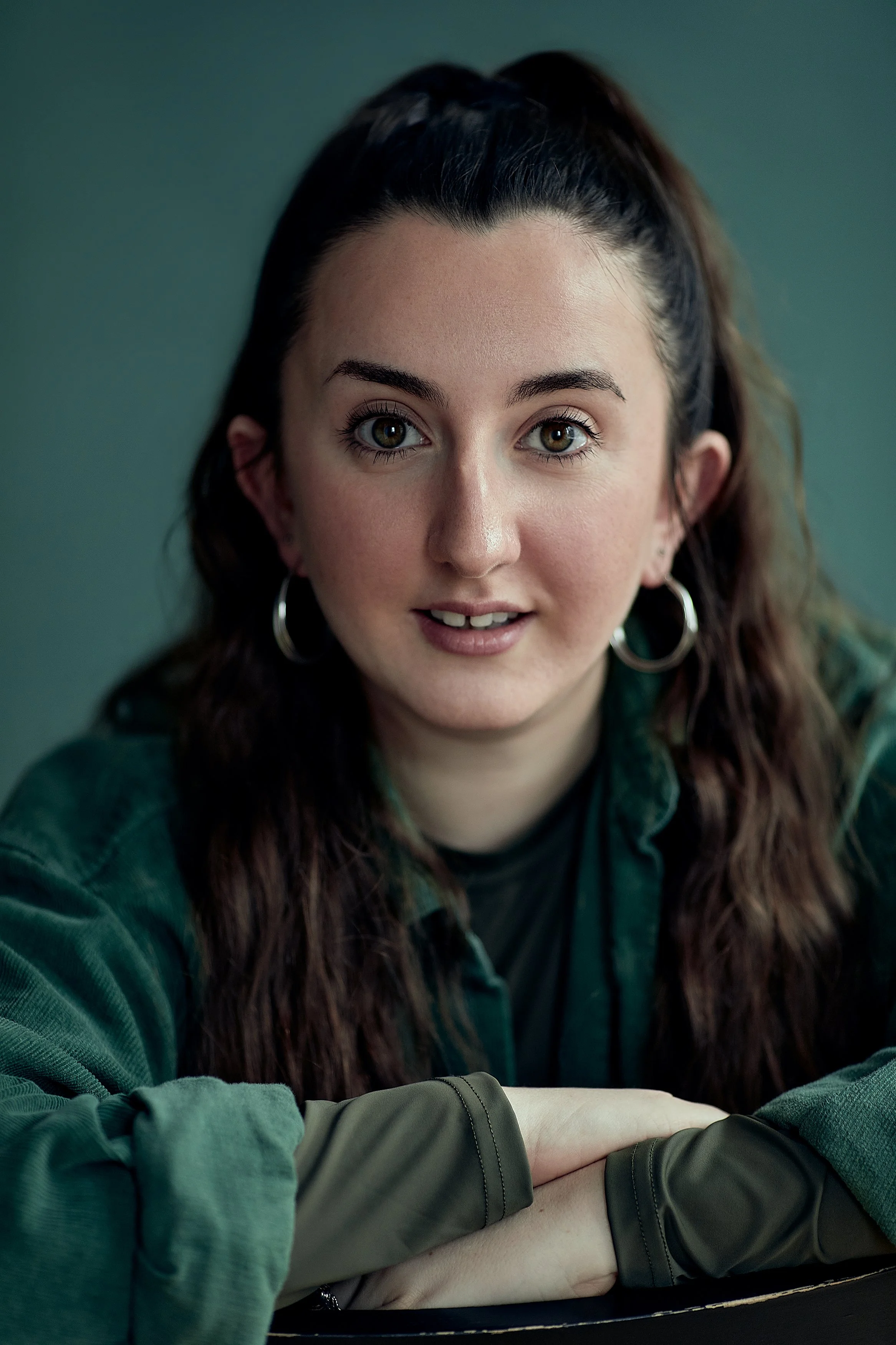 Close-up of a young woman with long wavy brown hair, hazel eyes, wearing hoop earrings and a green jacket, looking at the camera with a slight smile.