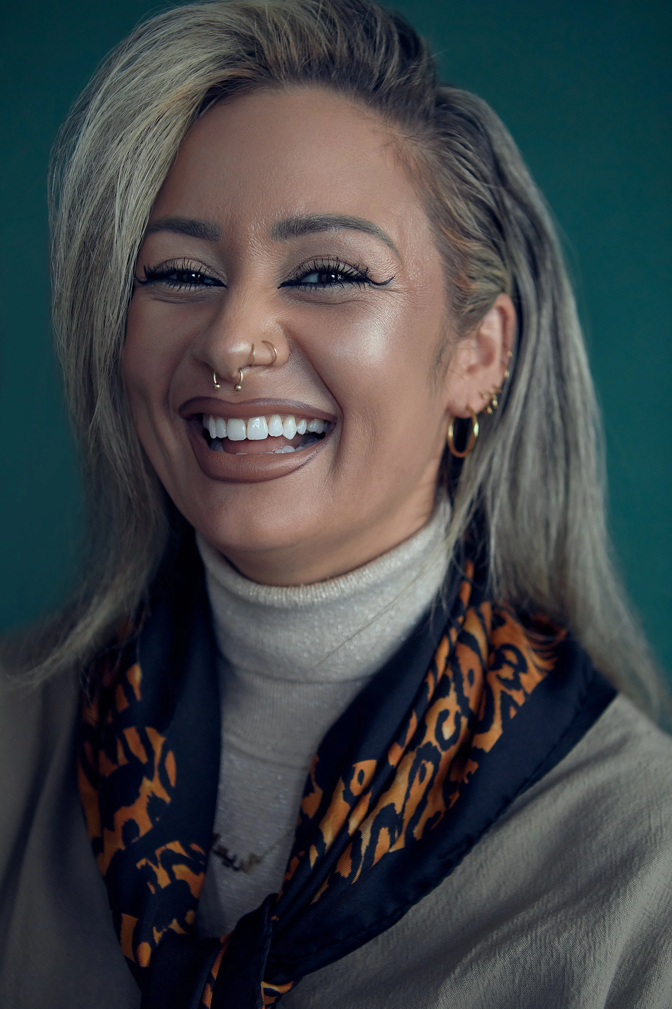 A woman with blonde and gray hair, multiple facial piercings, gold earrings, and makeup, smiling and showing her teeth, wearing a beige turtleneck sweater and a leopard print scarf.