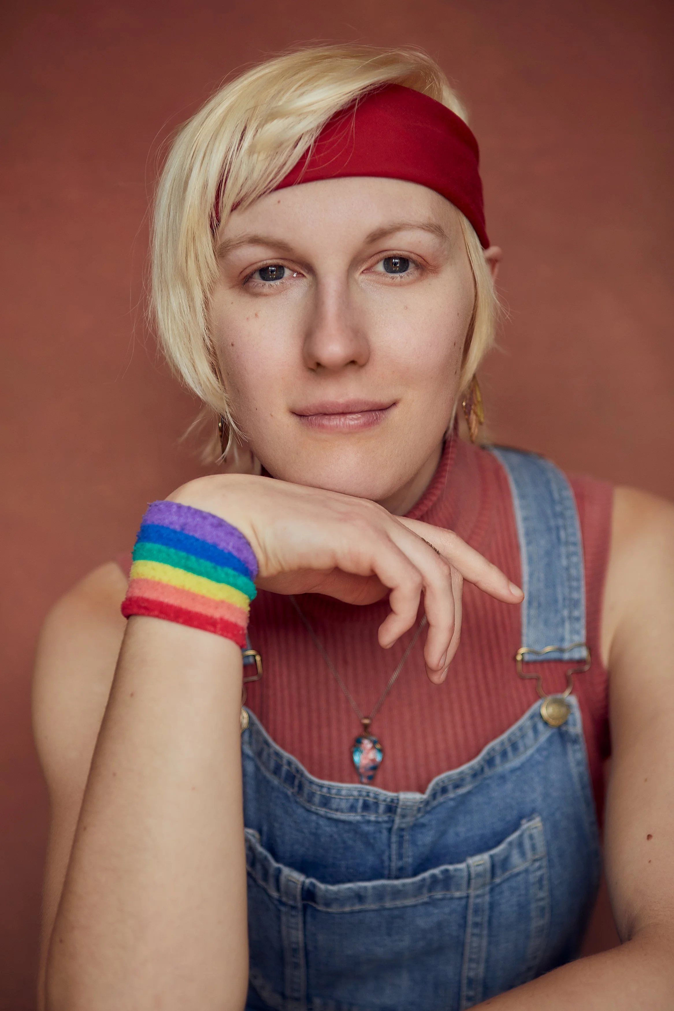 A young woman with short blonde hair wearing a red headband, rainbow wristbands, a red top, denim overalls, and jewelry, resting her chin on her hand against a pink background.