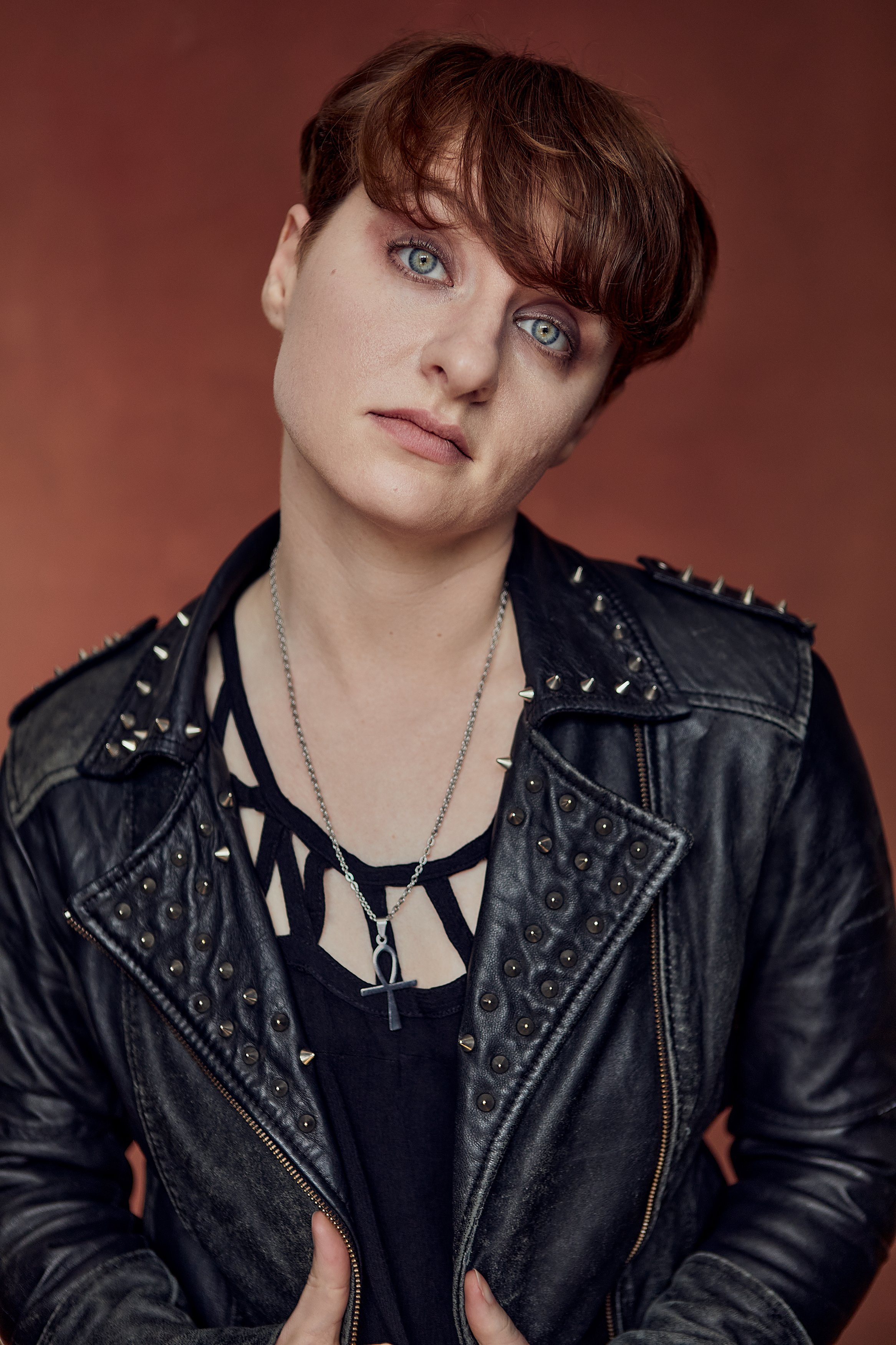 A person with short brown hair, blue eyes, and fair skin wearing a black leather jacket with silver studs, a black layered shirt, and chain necklaces, standing against a reddish-brown background.
