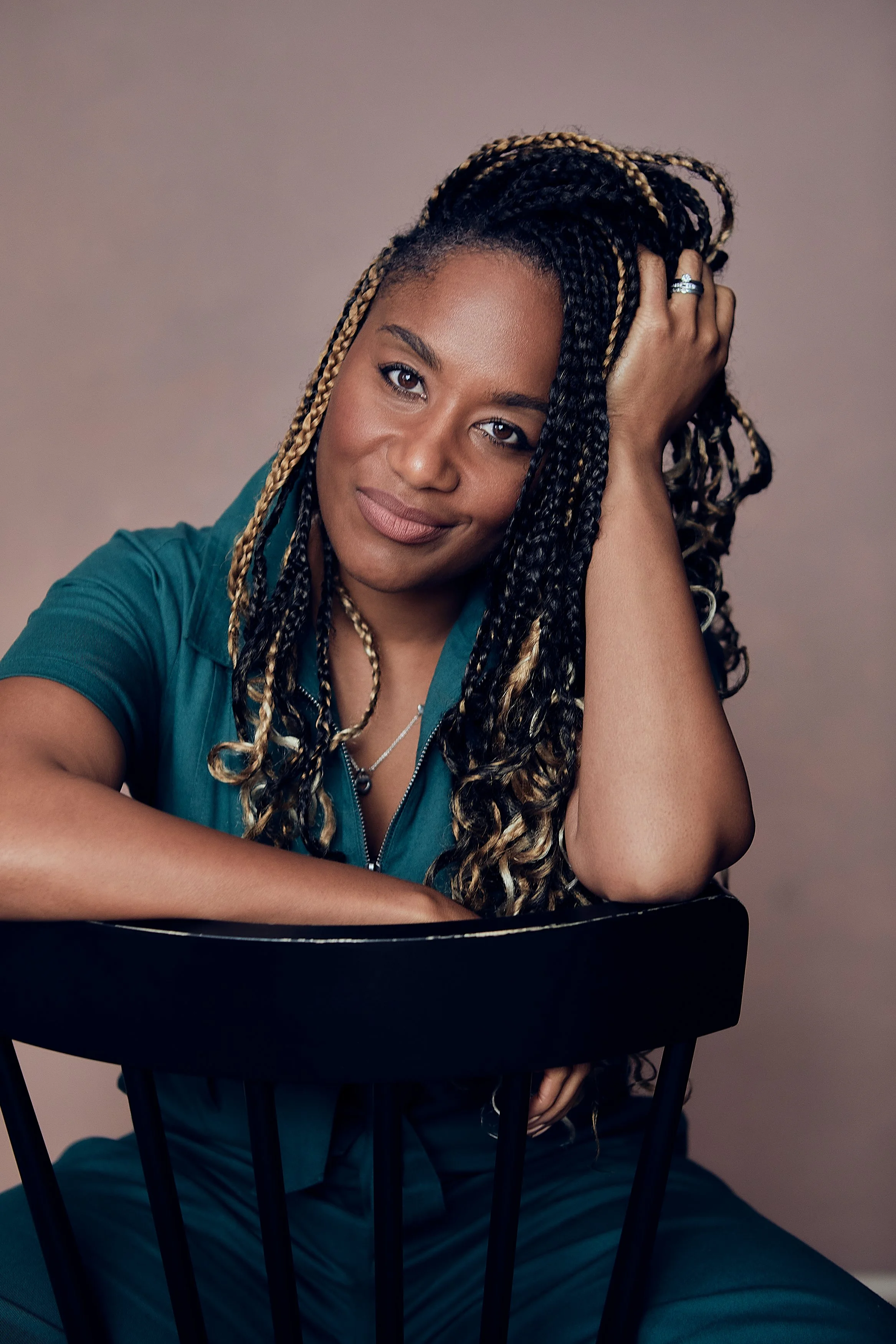 A woman with dark skin and long braided hair with blonde highlights sitting on a black chair, resting her arm on the chair's backrest, smiling softly at the camera, with a beige background.