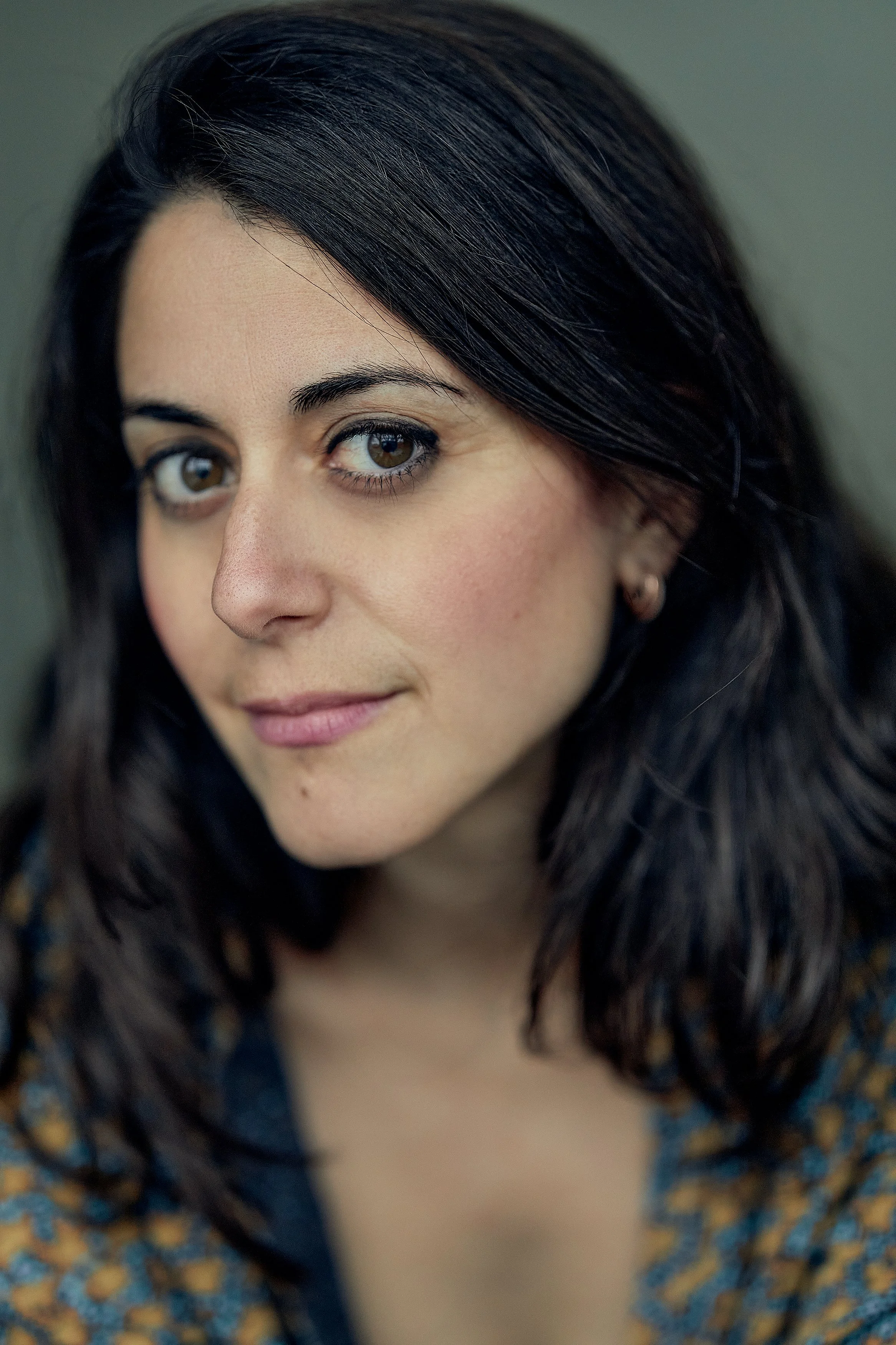 Close-up portrait of a woman with dark brown hair, brown eyes, and light skin, wearing a floral top and small earrings.