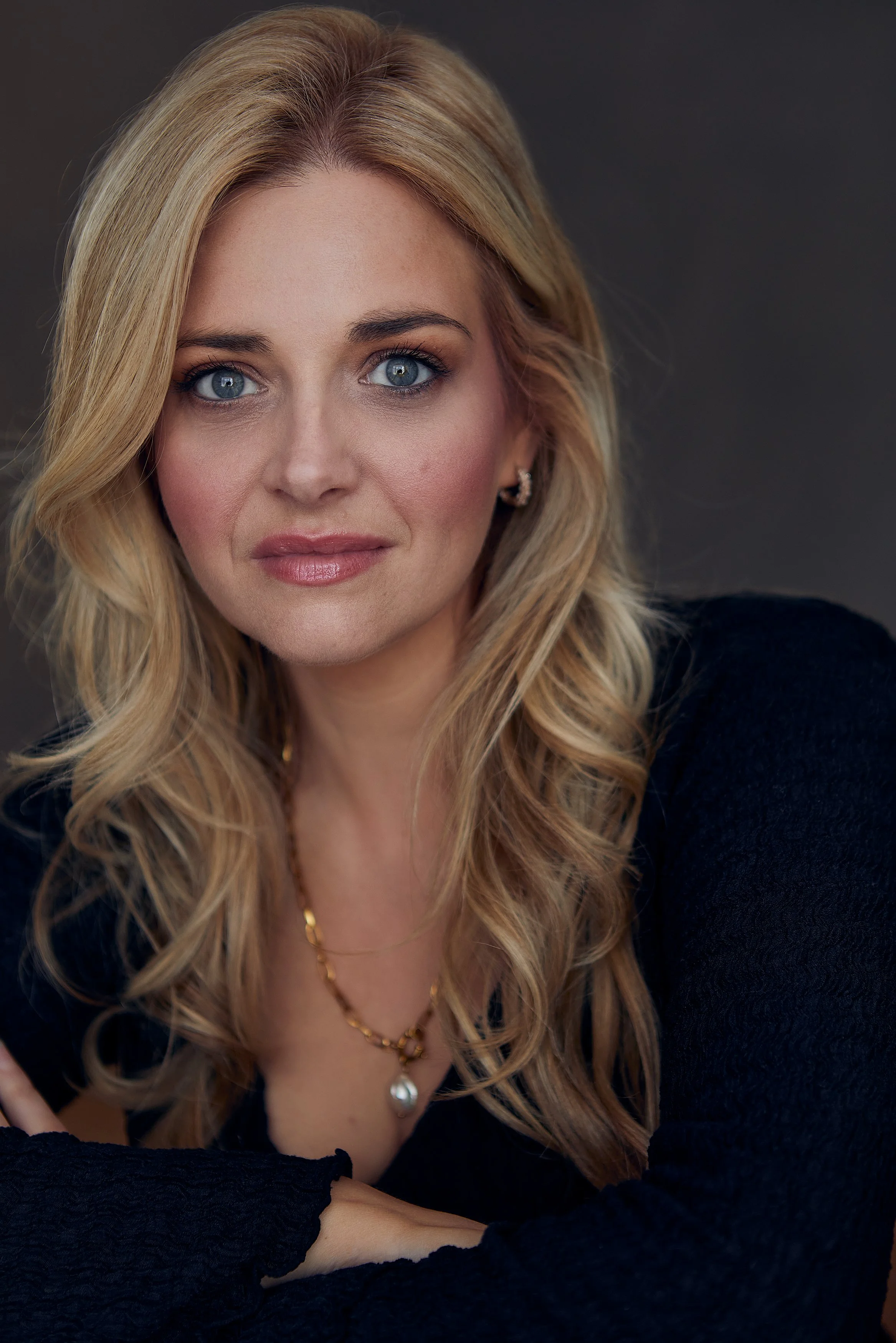 A close-up portrait of a woman with long blonde hair, blue eyes, and wearing a black top, gold jewelry, and earrings, against a dark background.