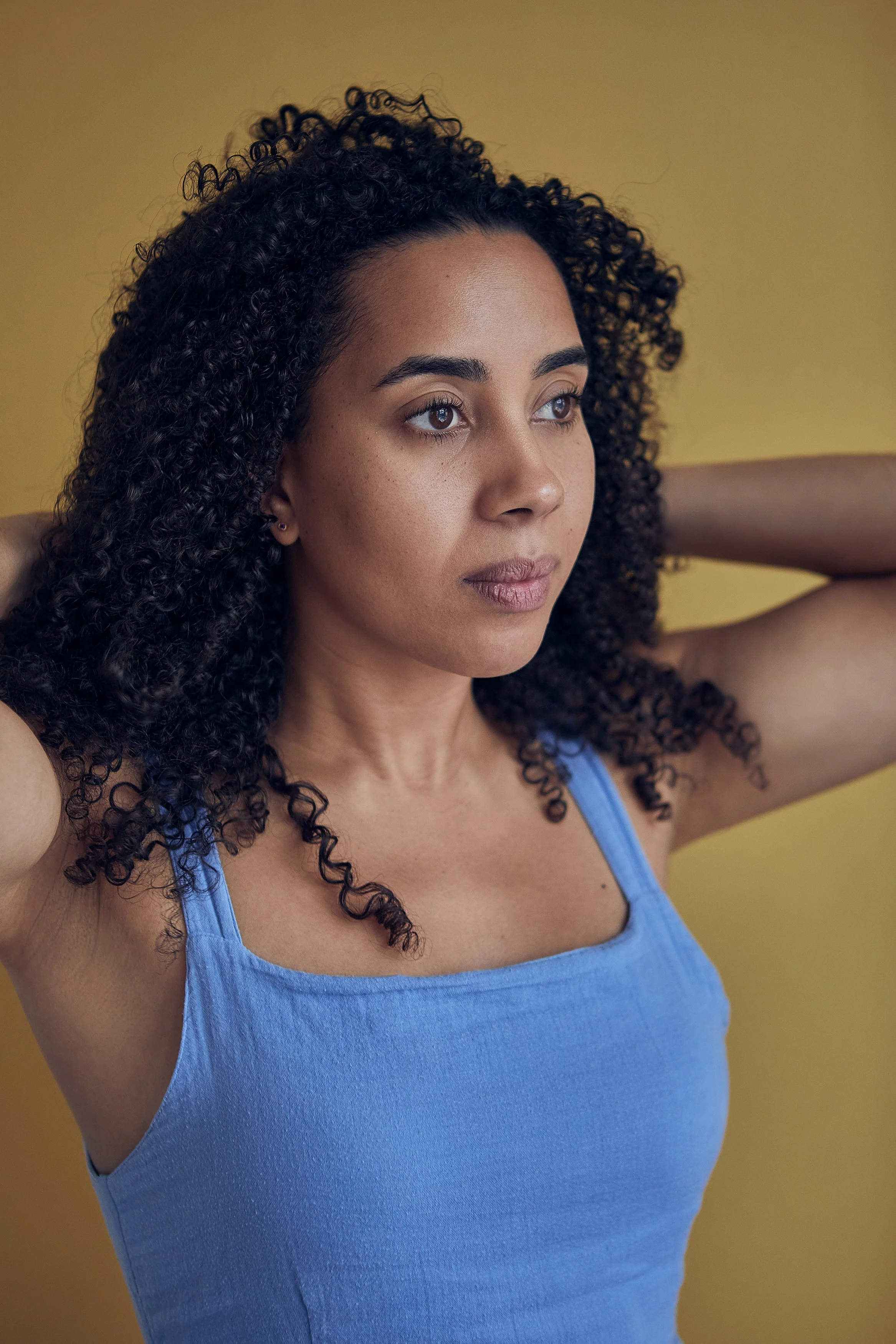 A woman with curly dark hair and medium skin tone wearing a blue sleeveless top, standing against a yellow background, with her arms raised behind her head.