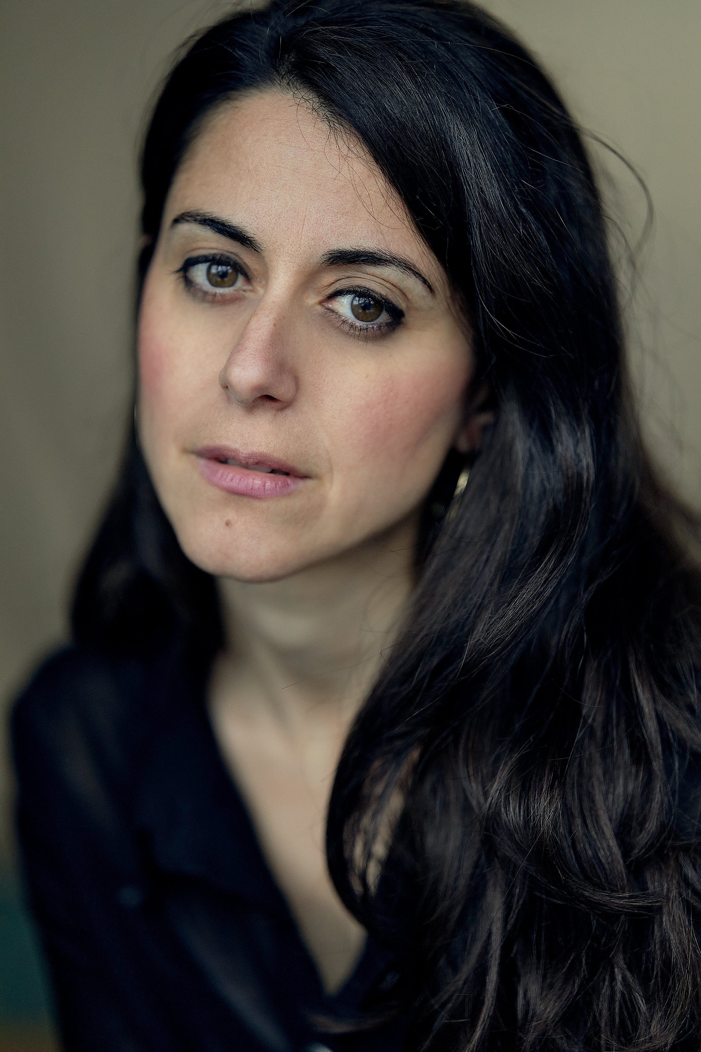 Close-up portrait of a woman with long dark hair, wearing makeup, a black top, and gold hoop earrings, looking at the camera.
