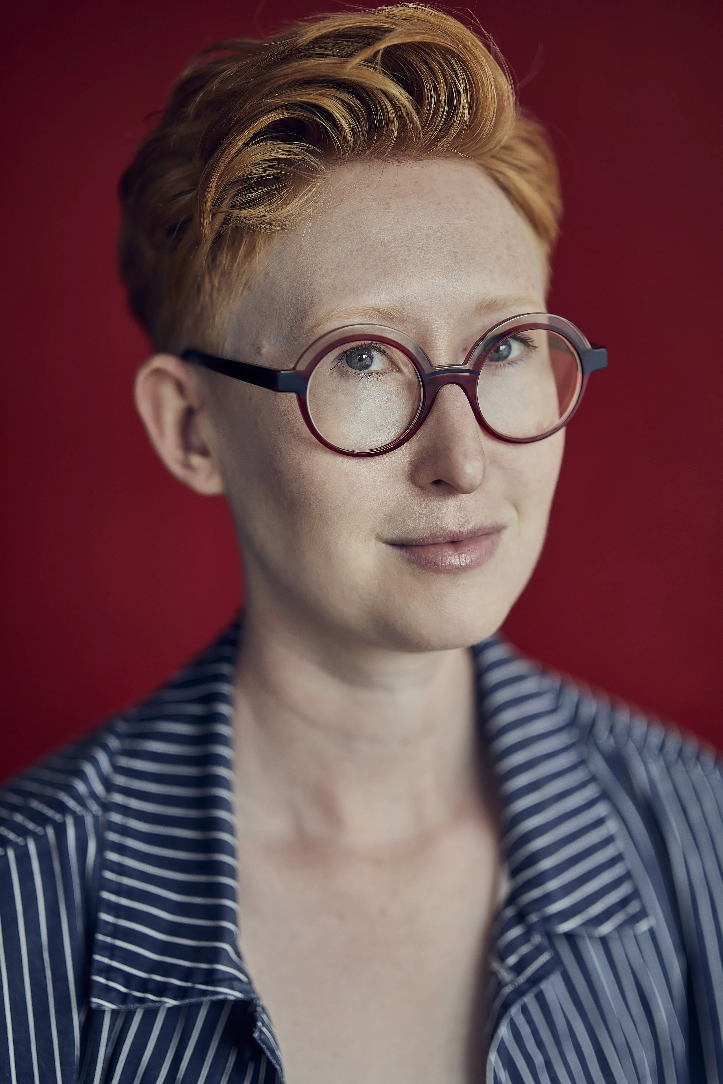 A woman with short red hair, wearing round glasses with dark red frames, and a blue and white striped shirt, standing against a red background.