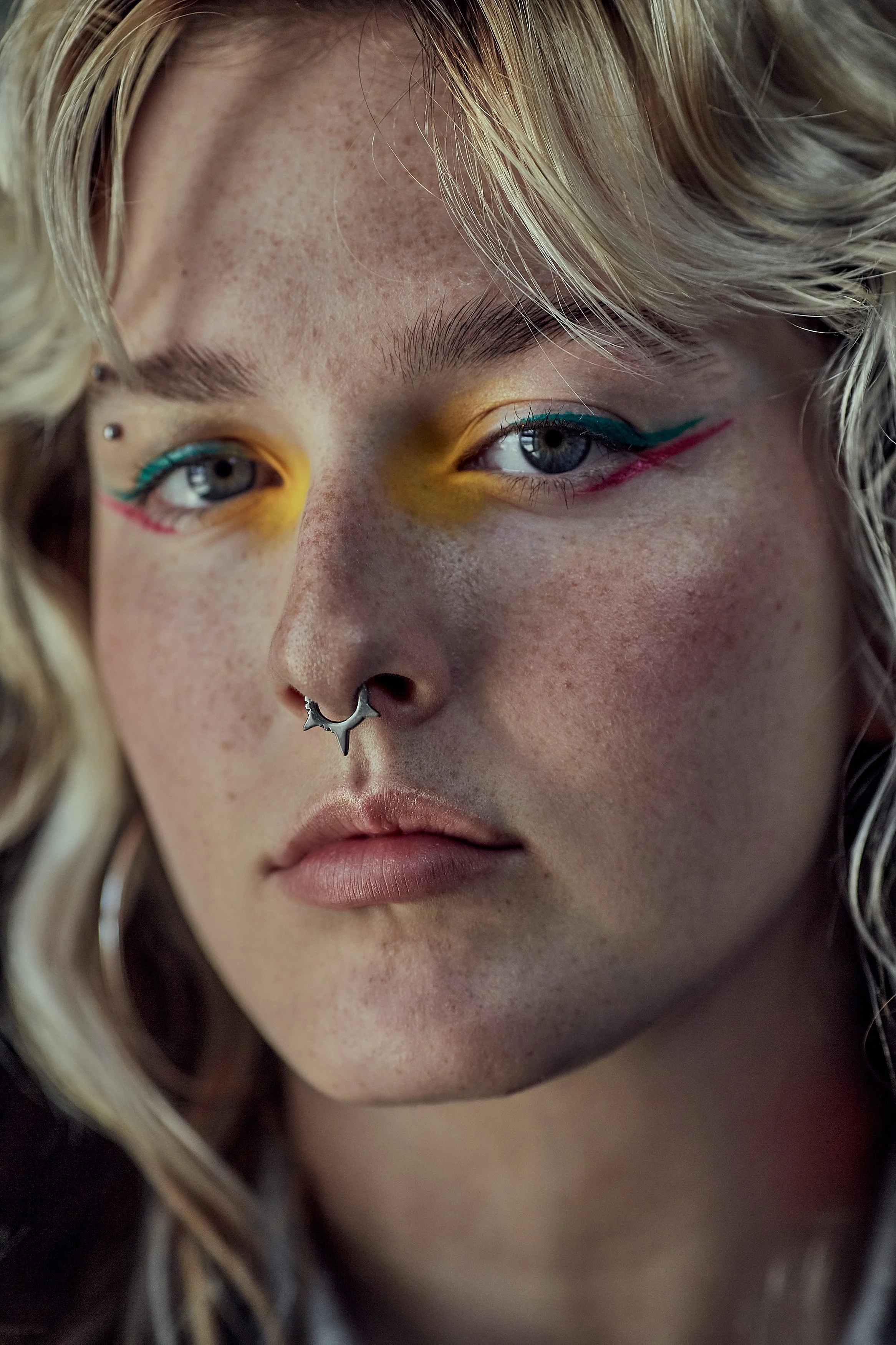 Close-up of a person with fair skin, freckles, platinum blonde wavy hair, wearing colorful makeup with yellow eyeshadow and teal and pink eyeliner, and a septum piercing.