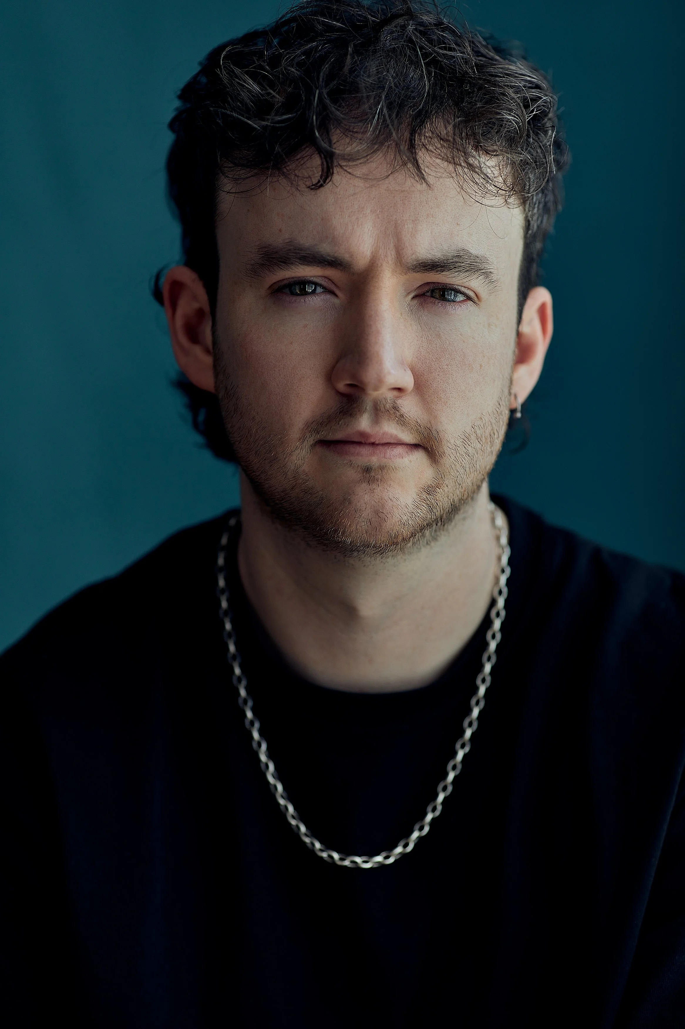 Close-up of a young man with curly dark hair, light skin, and blue eyes, wearing a silver chain necklace and a black shirt, against a dark teal background.