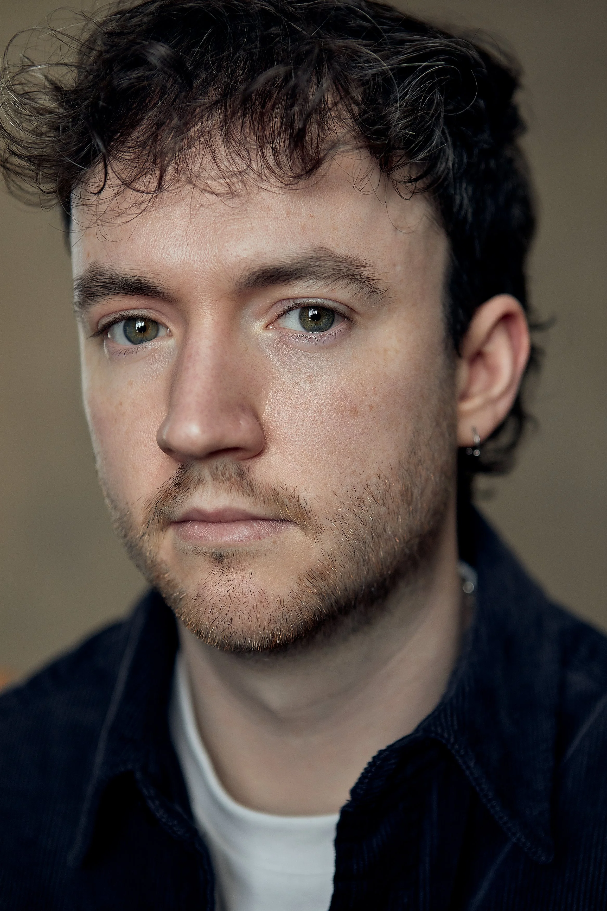Close-up portrait of a young man with curly dark hair, light skin, and green eyes, wearing a dark jacket over a white shirt, looking slightly to the side.