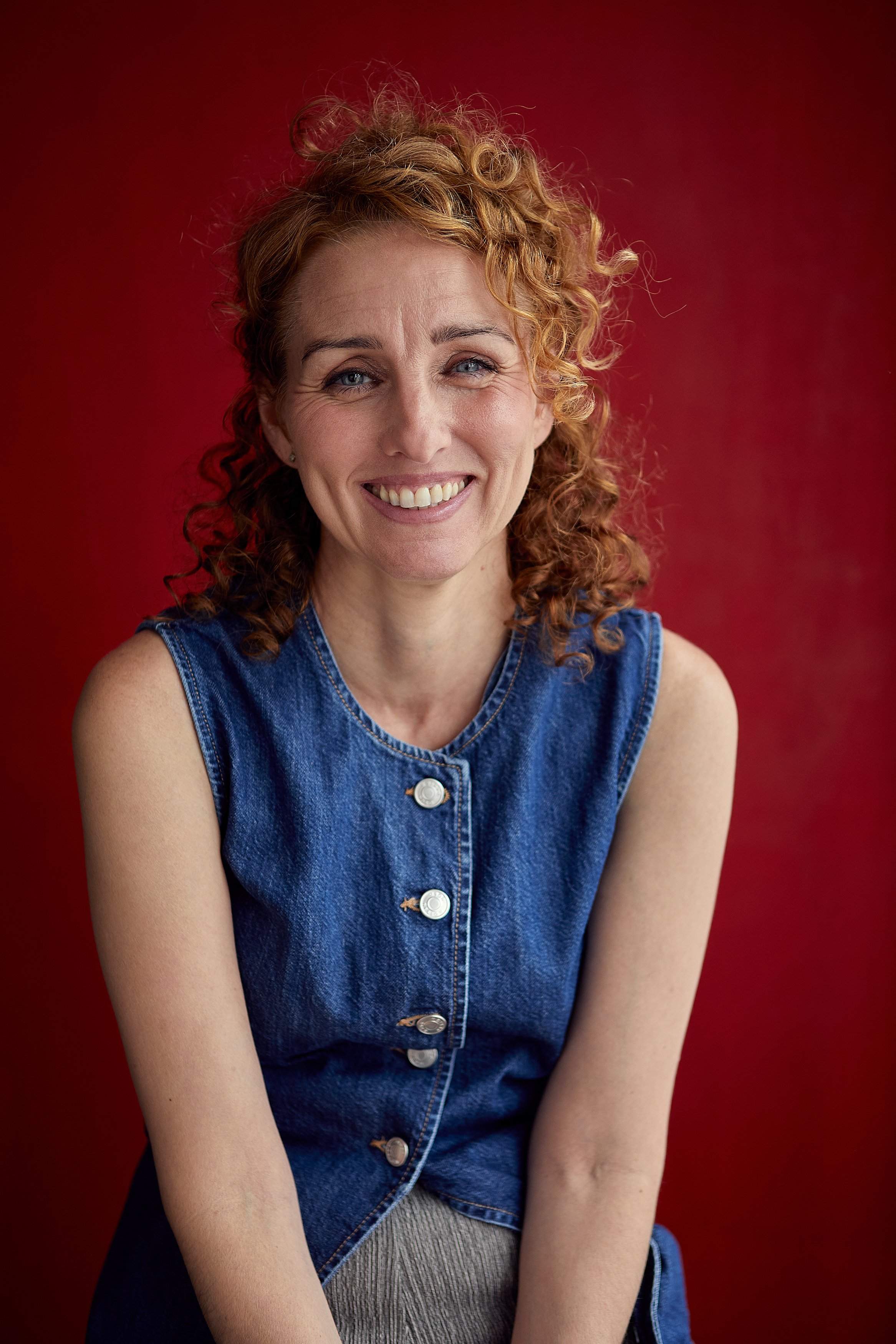 A woman with curly red hair smiling, wearing a sleeveless denim top, against a red background.