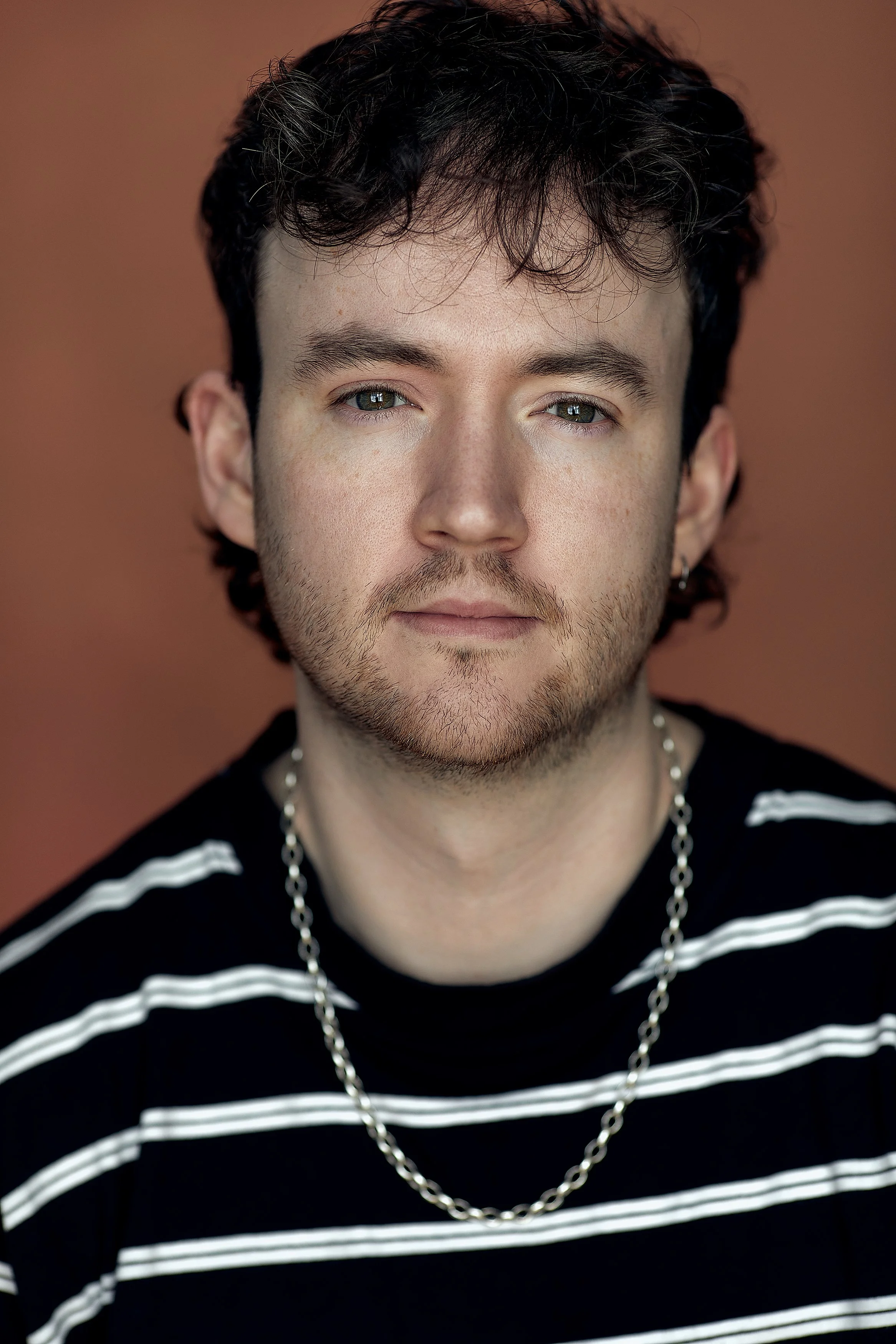 A young man with dark, curly hair, light skin, and blue eyes, wearing a black and white striped shirt, a silver chain necklace, and an earring, looks directly at the camera with a neutral expression.